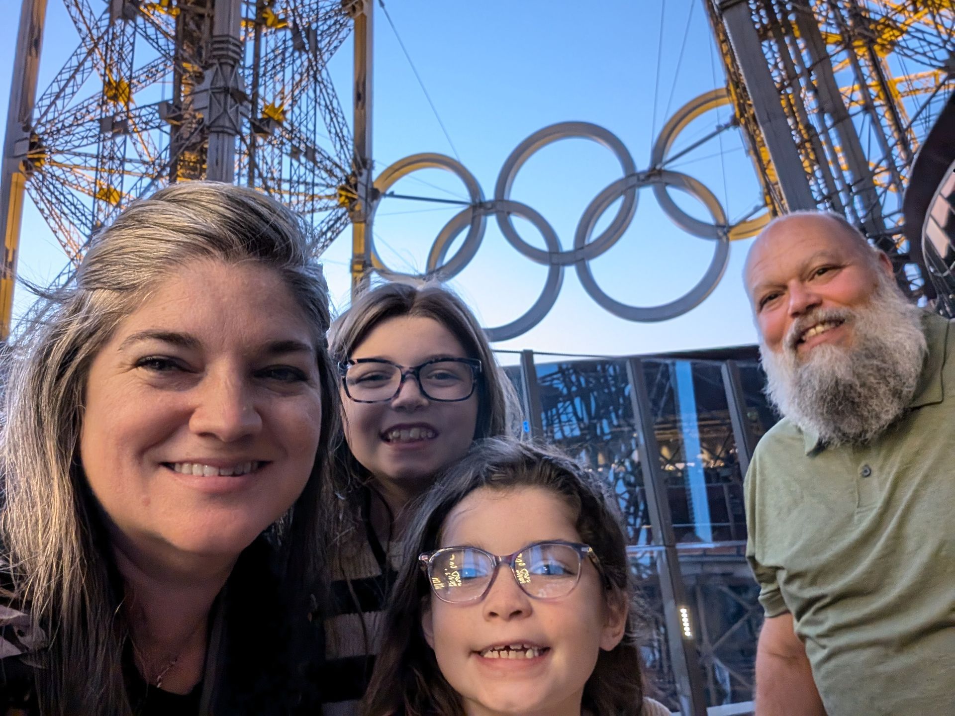 Family selfie in front of the Eiffel Tower and Olympic rings; smiles, outdoor setting.