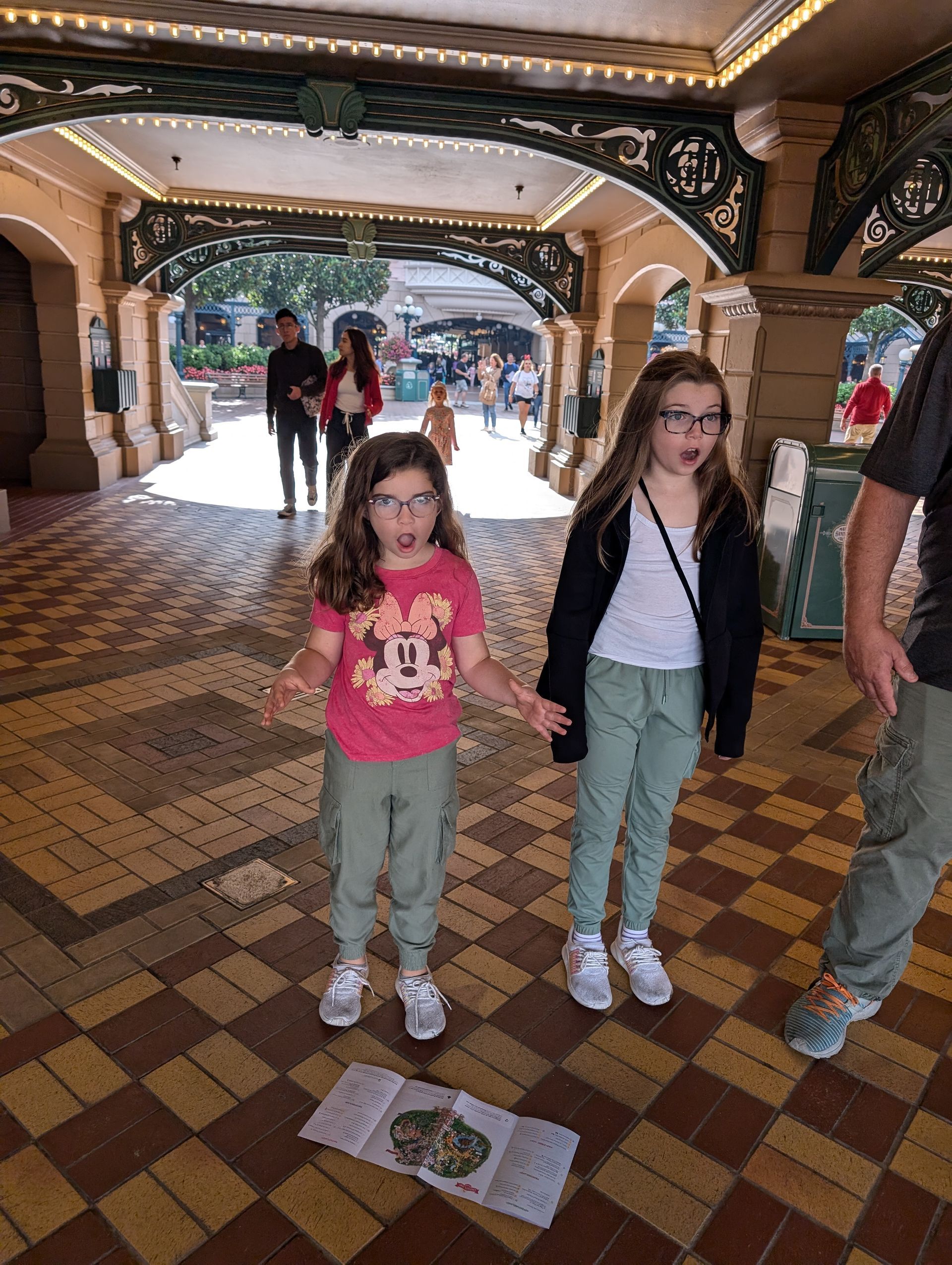 Two girls with glasses looking at something on the ground, in an archway with people in the background.