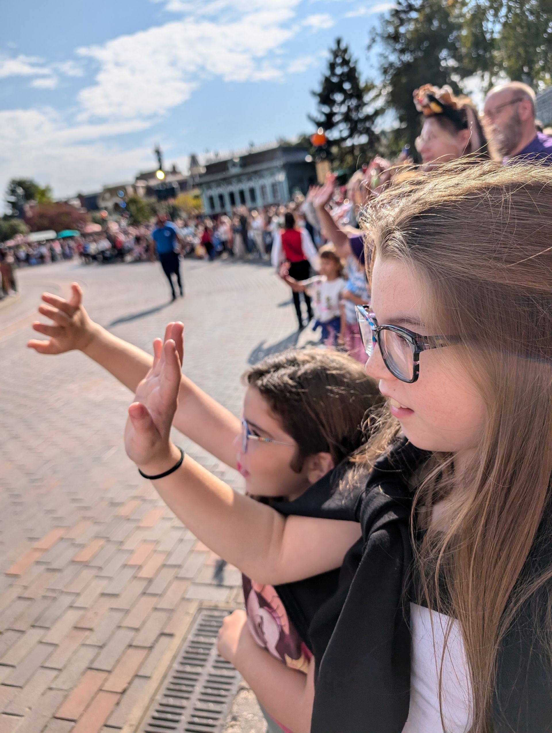 Two girls watching a parade, waving their arms with a crowd in the background. Sunny day.