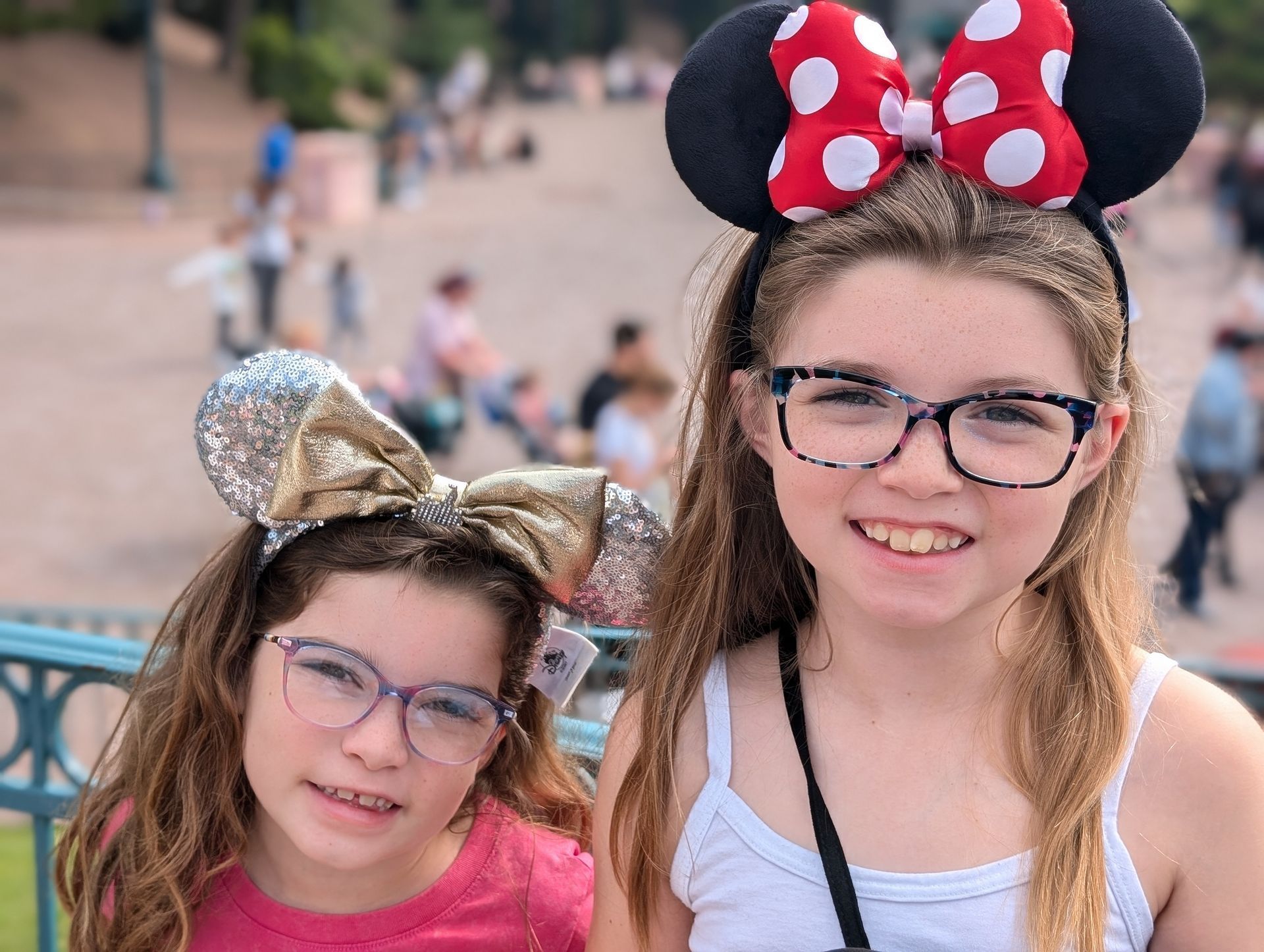 Two girls with Minnie Mouse ears, smiling in front of a blurred outdoor scene.