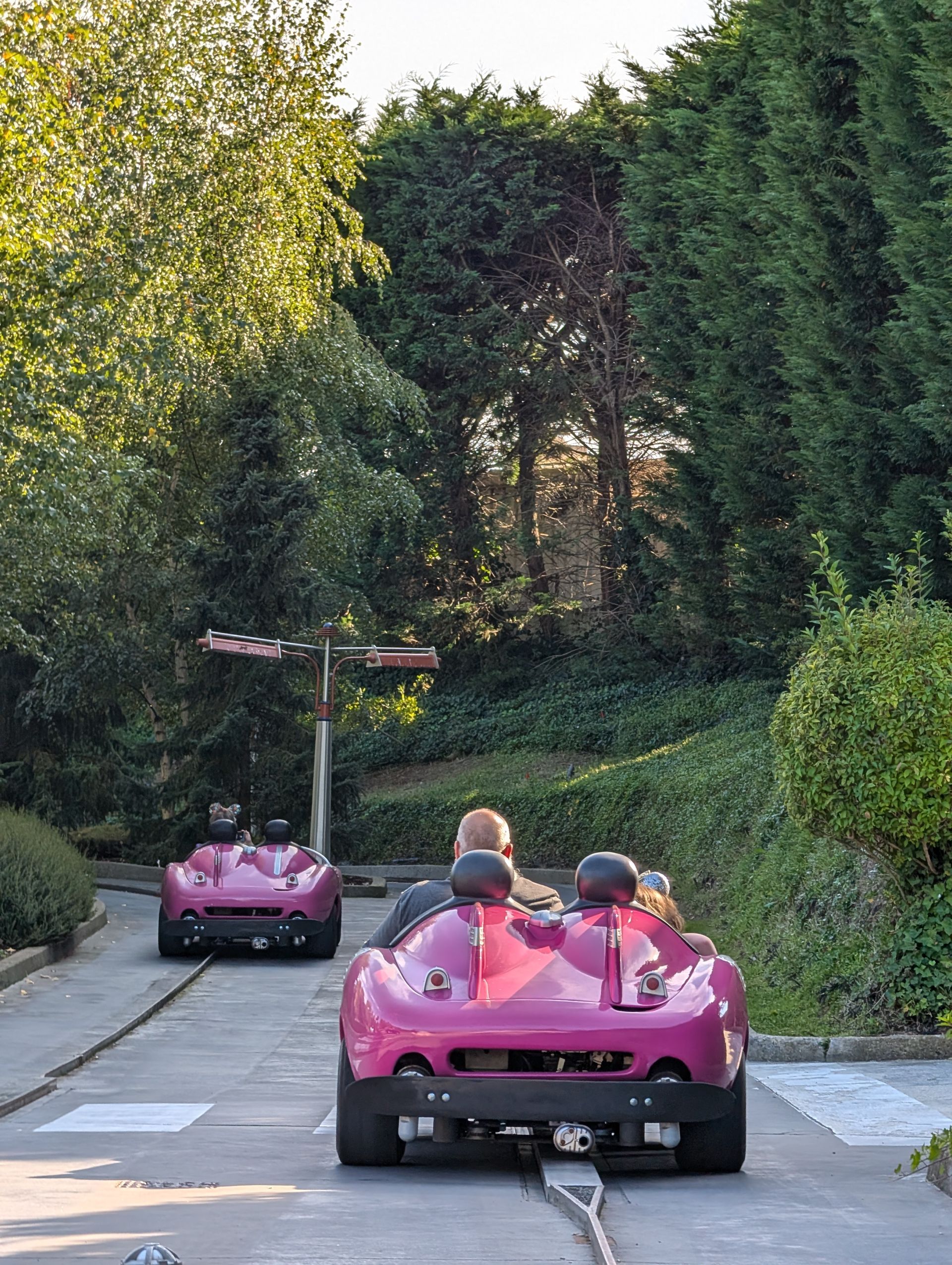 Two pink amusement park cars on a track with passengers, trees in the background.