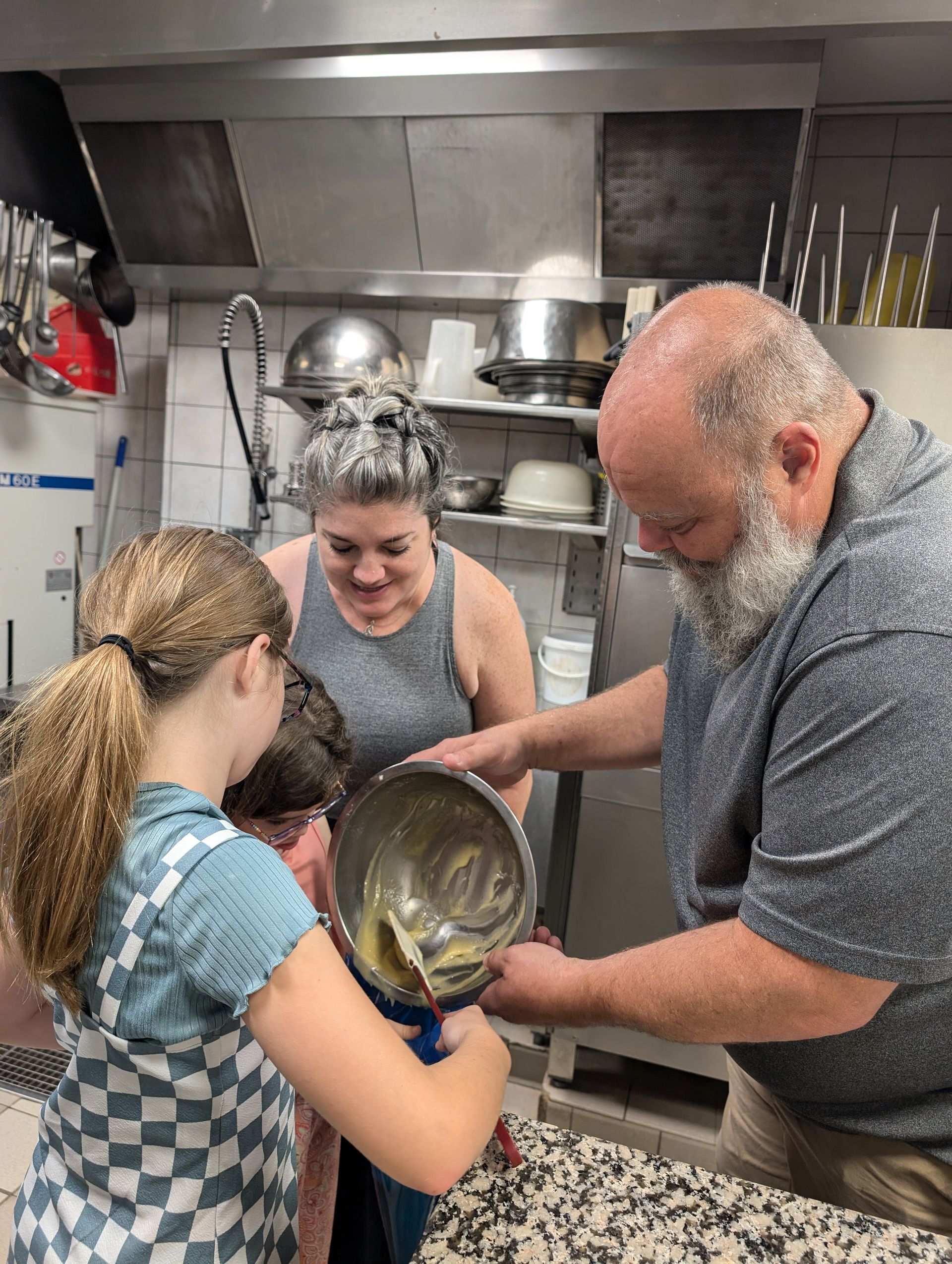 Person pouring batter from a bowl into a bowl held by a child, watched by another person, in a kitchen.