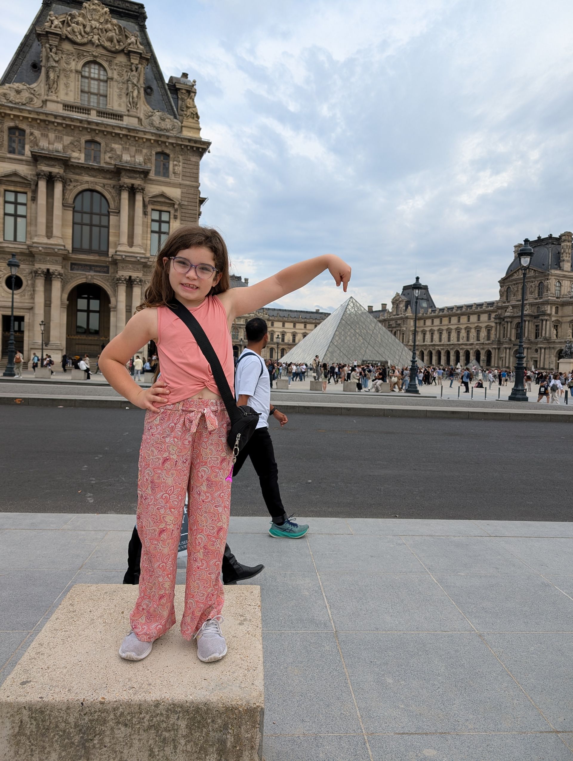 Girl in floral pants poses in front of the Louvre Museum and pyramid, Paris.