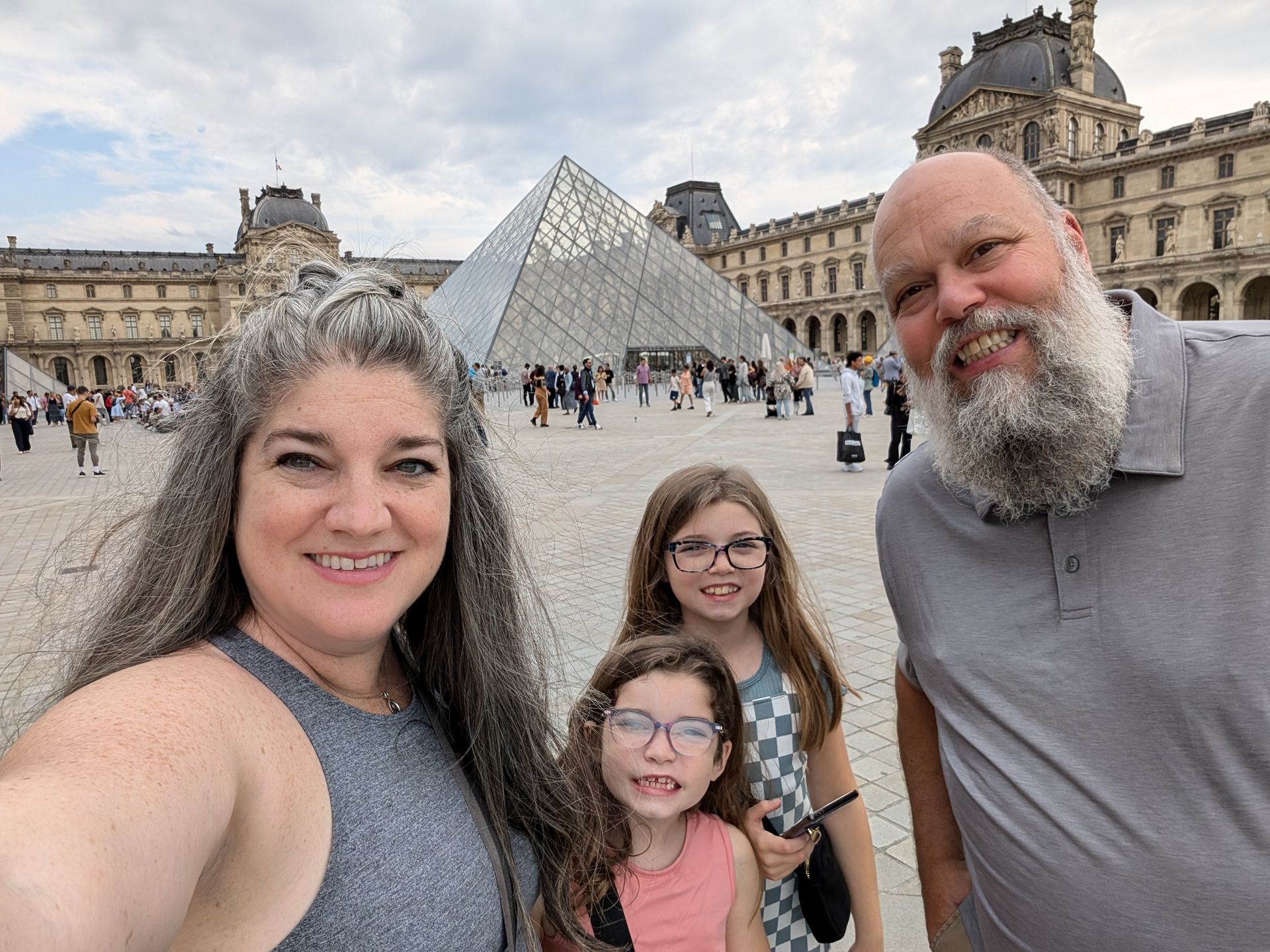Family selfie in front of the Louvre Museum in Paris, with pyramid and building visible.