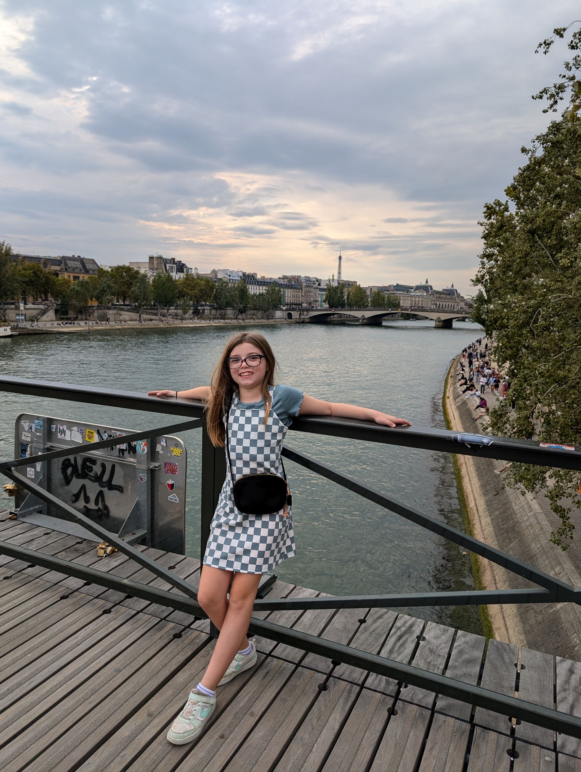 Girl on a bridge over a river, posing. Wearing a checkered dress, smiling. City buildings in the background.