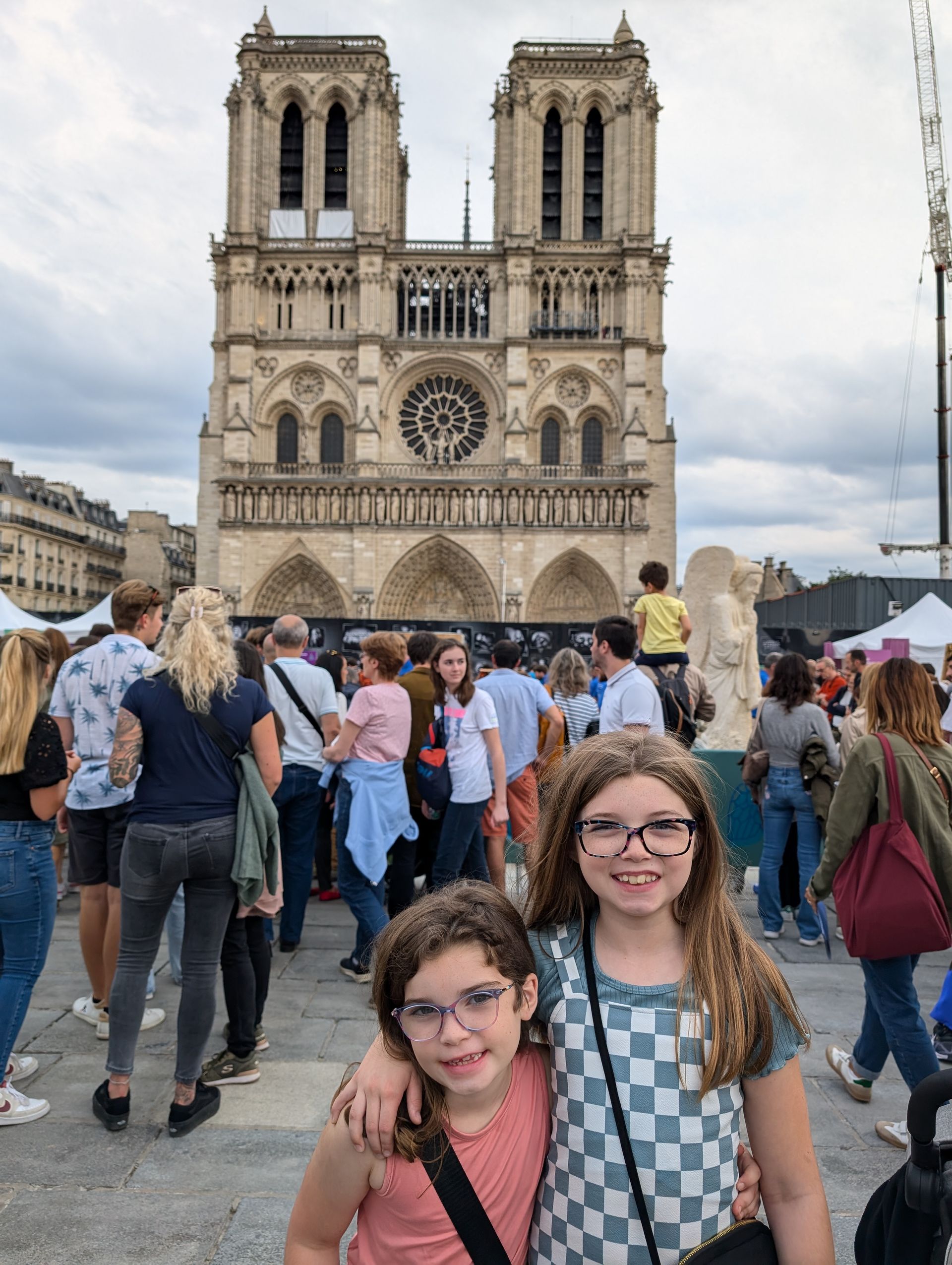 Two girls pose in front of Notre Dame Cathedral, crowds of people in background.