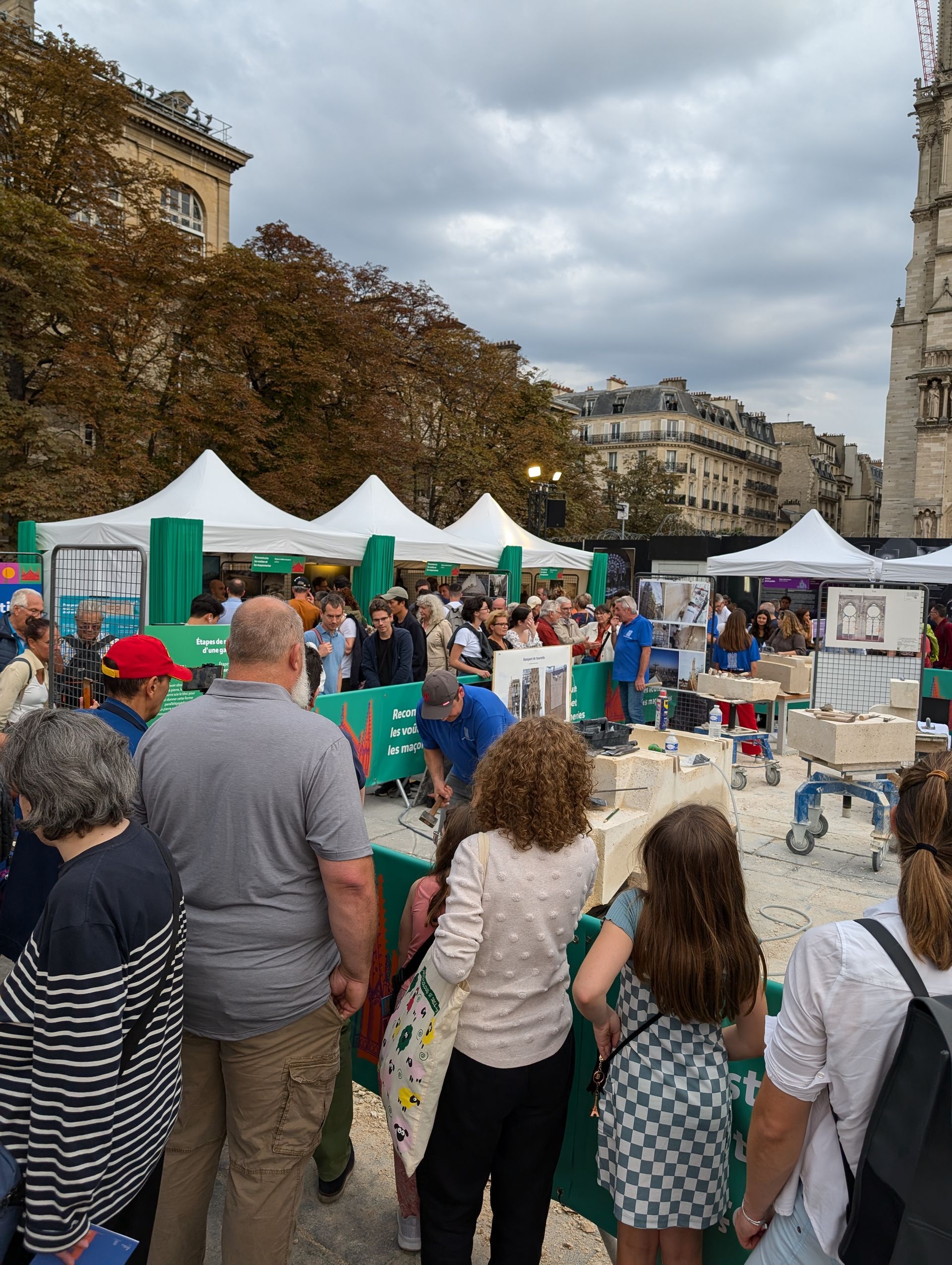 Outdoor market with tents and many people; buildings and trees in the background.