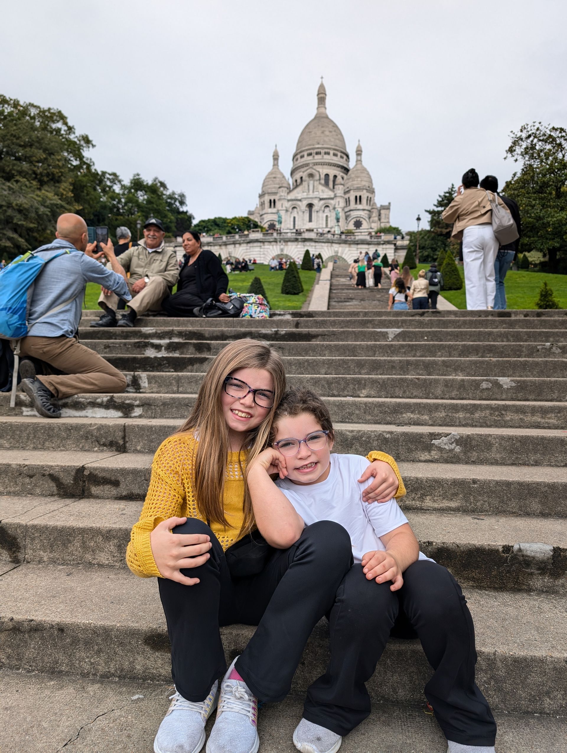 Two children sit on stone steps, smiling at the camera, with a white domed cathedral in the background.