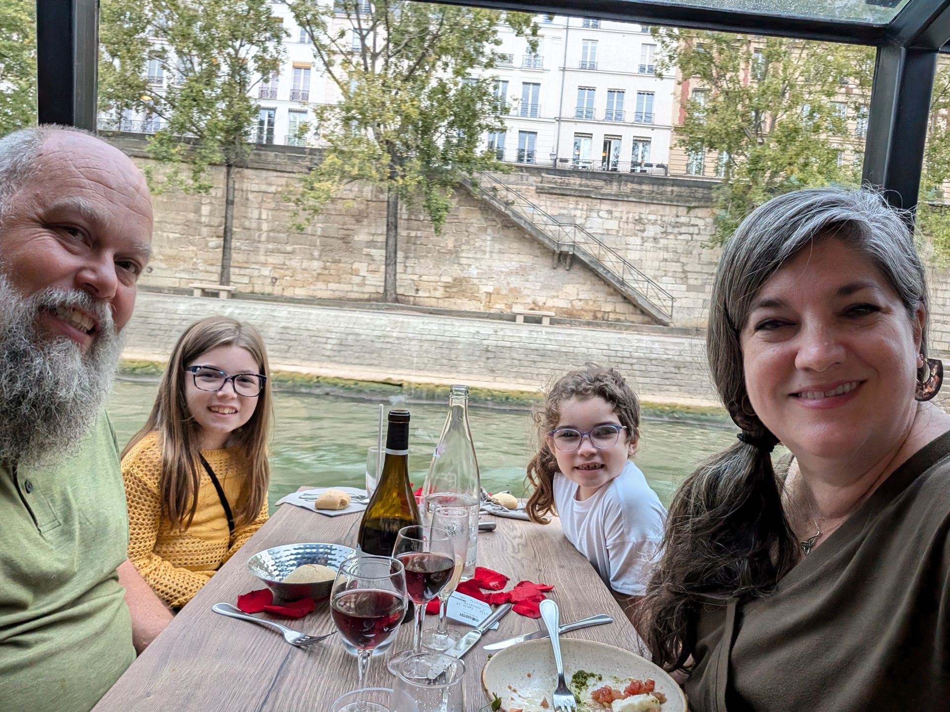 Family dining on a boat on a river, with buildings in the background. They are smiling, with food and wine on the table.