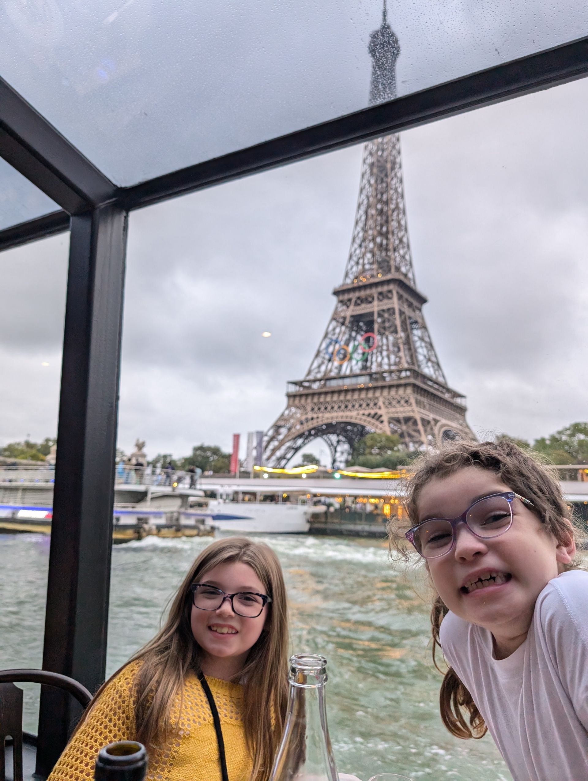 Two children on a boat in Paris, smiling in front of the Eiffel Tower on an overcast day.