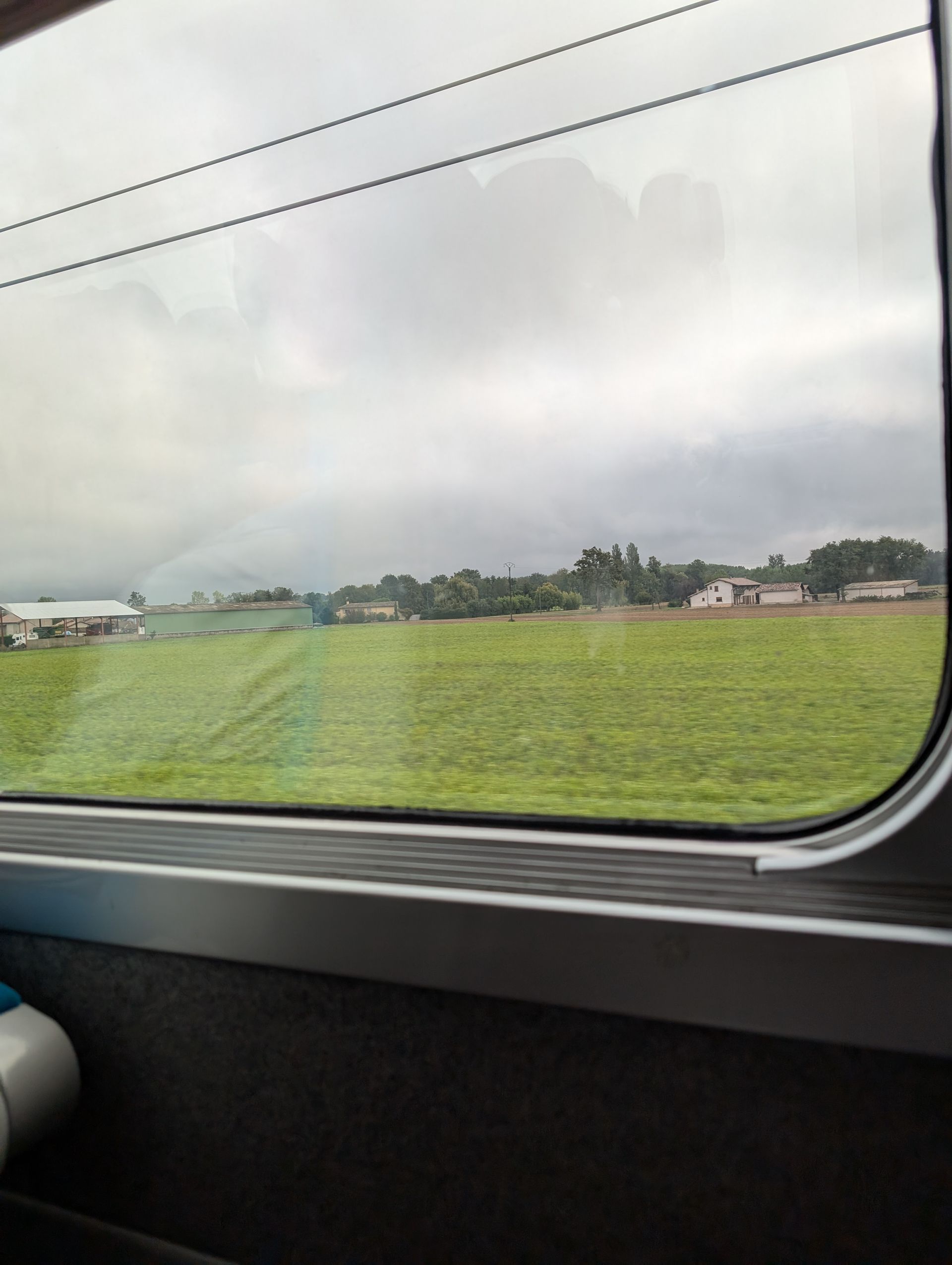 View from train window: green field, cloudy sky, buildings in the distance.