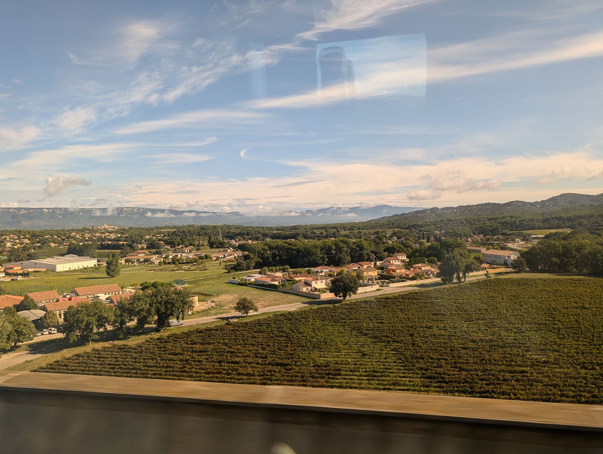 View from train window: green vineyard, houses, trees, and mountains under a blue sky with clouds.