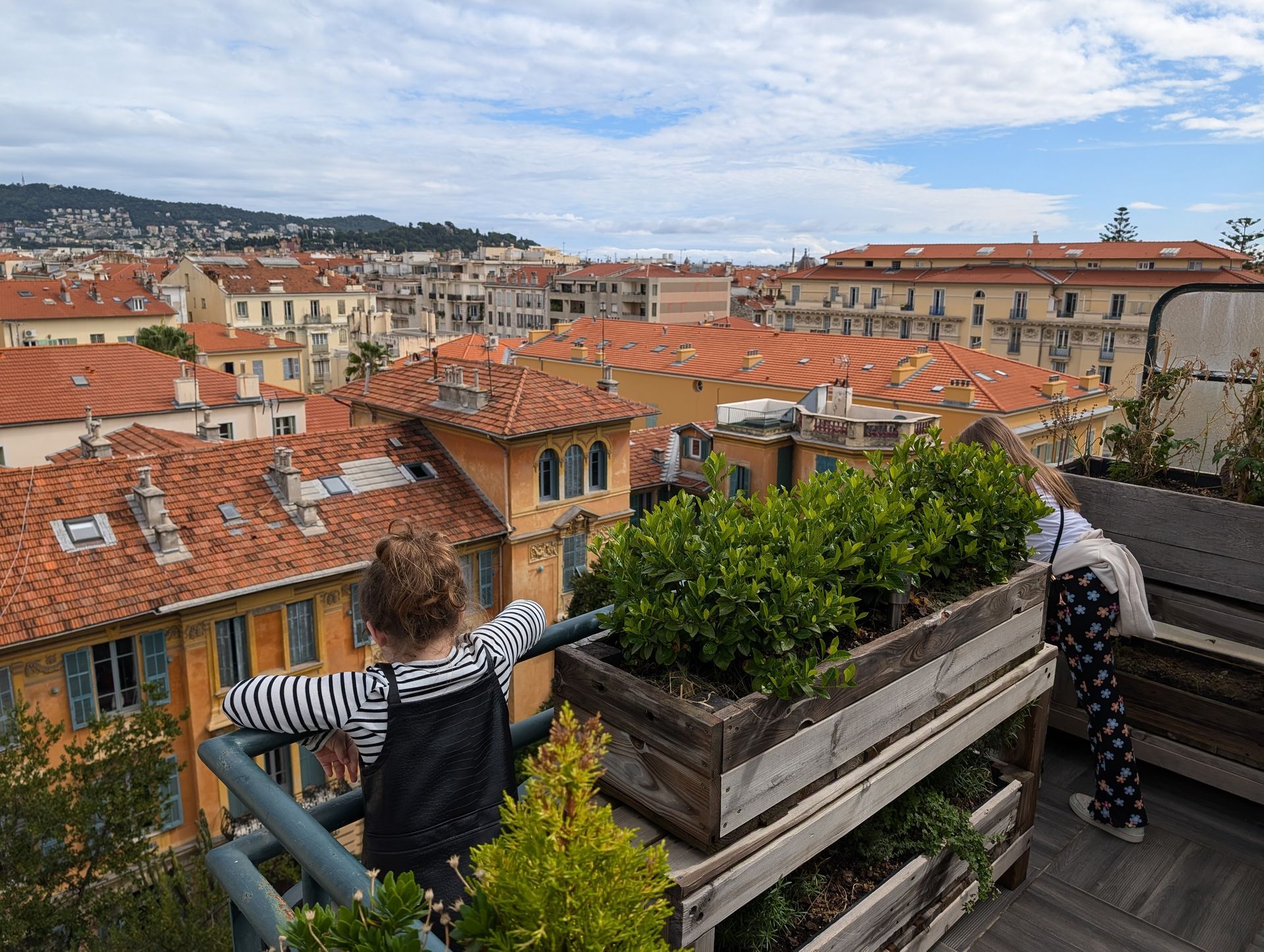 Children overlook a city with red tile roofs. Green plants and a balcony railing are in the foreground.