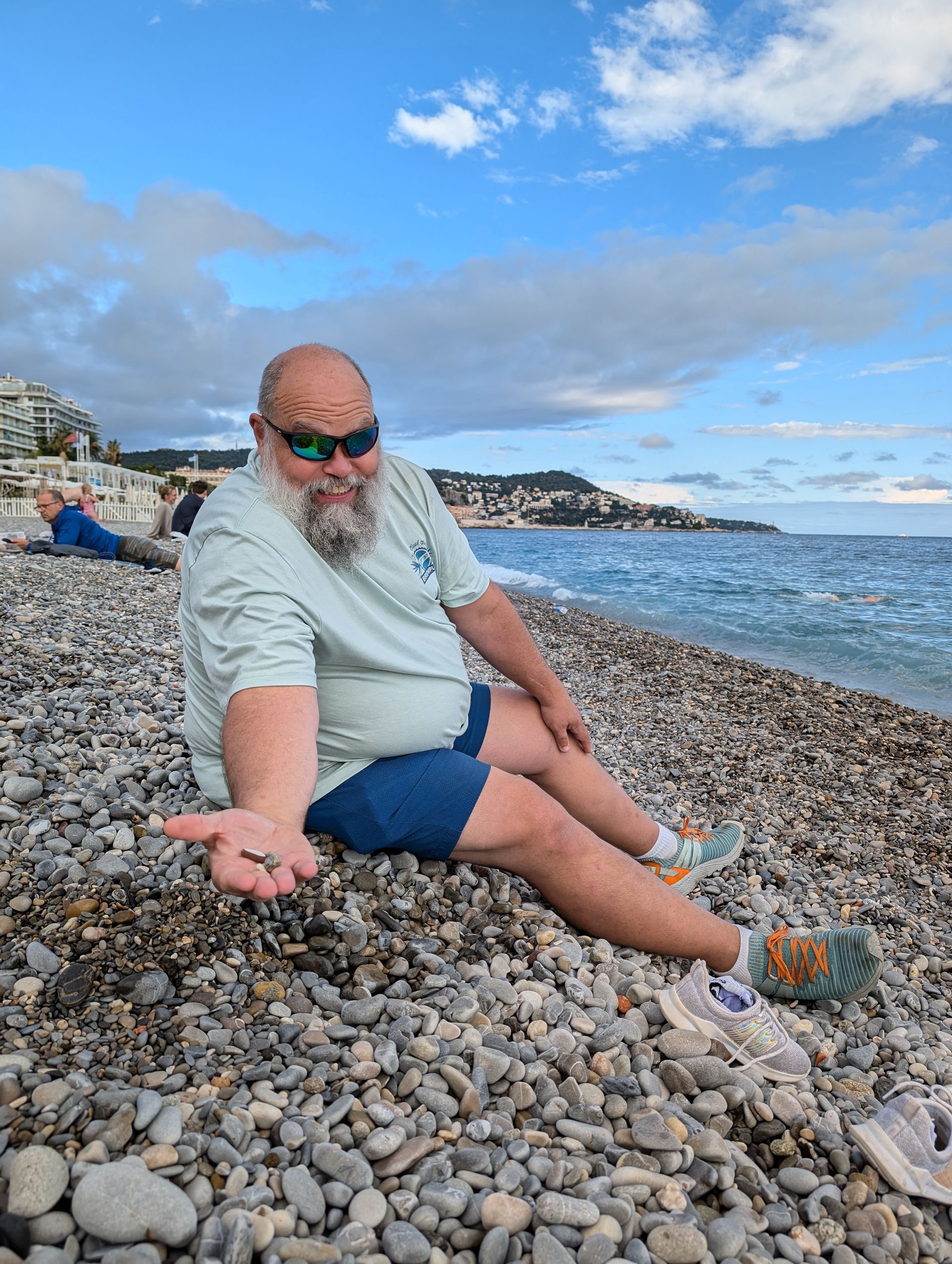 Man with beard holding pebbles on a pebbled beach. Blue sea and sky in background.