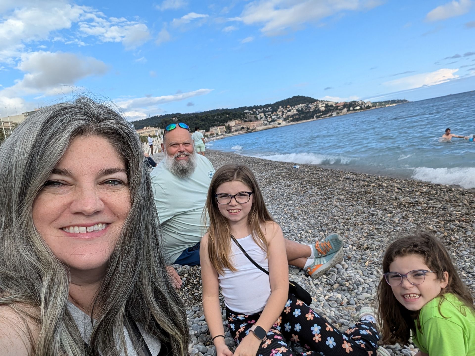 Family of four on a pebble beach, smiling. Ocean and coastal town in the background. Sunny day.