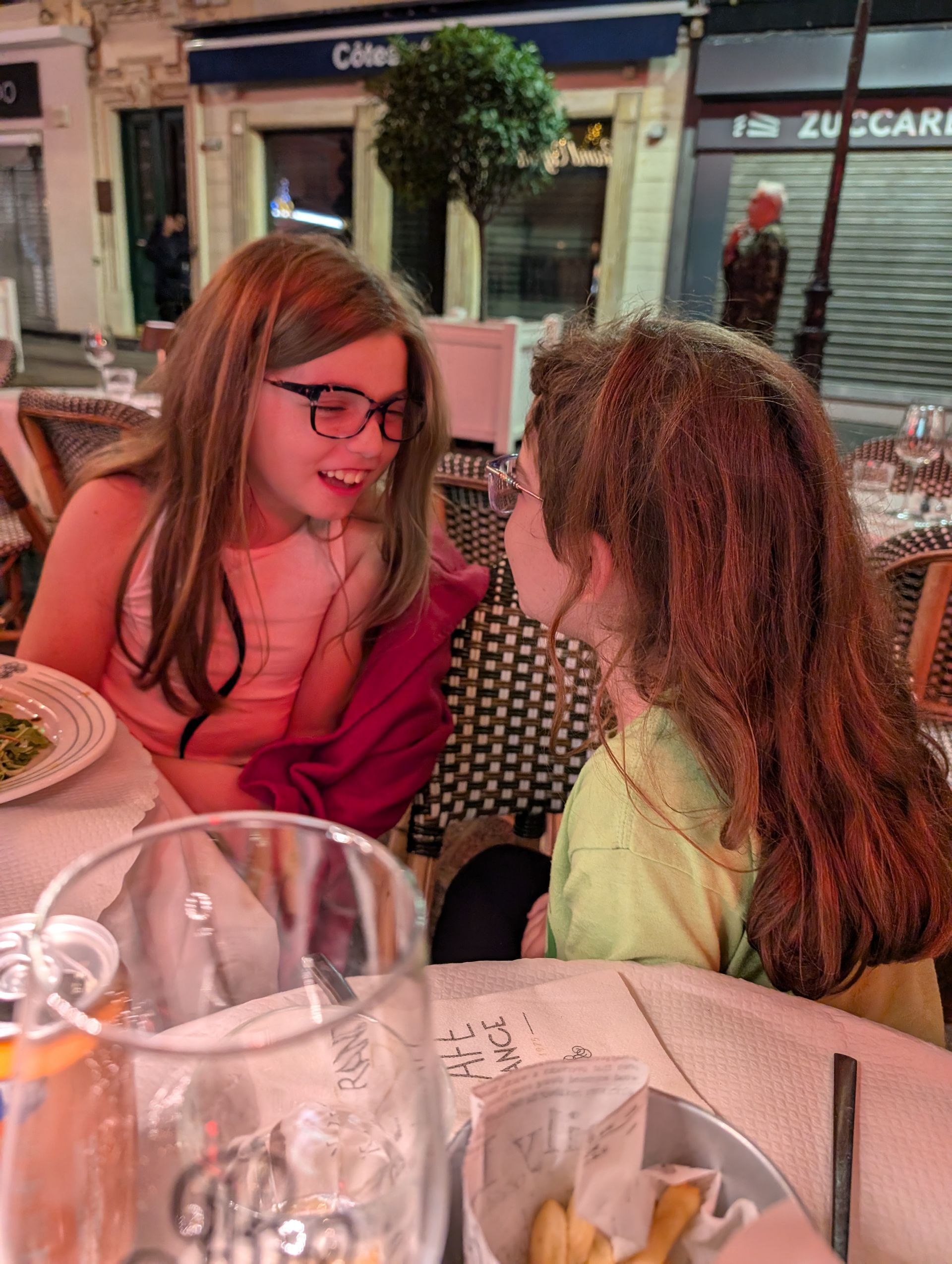 Two girls laughing at an outdoor cafe. One wears glasses and pink, the other has long hair and green. Paris.