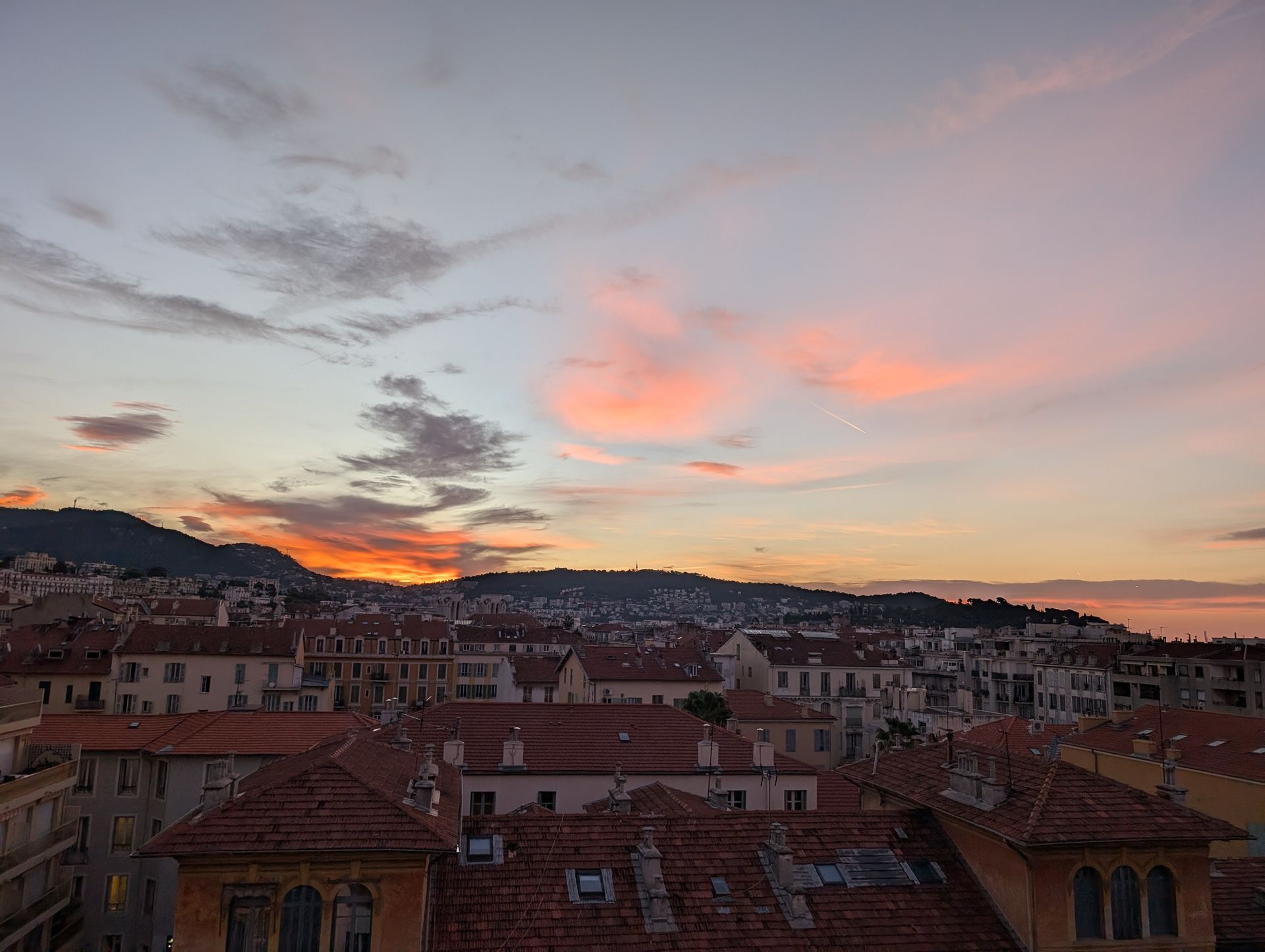 Sunset over a city with red-tiled roofs, mountains, and colorful clouds in the sky.