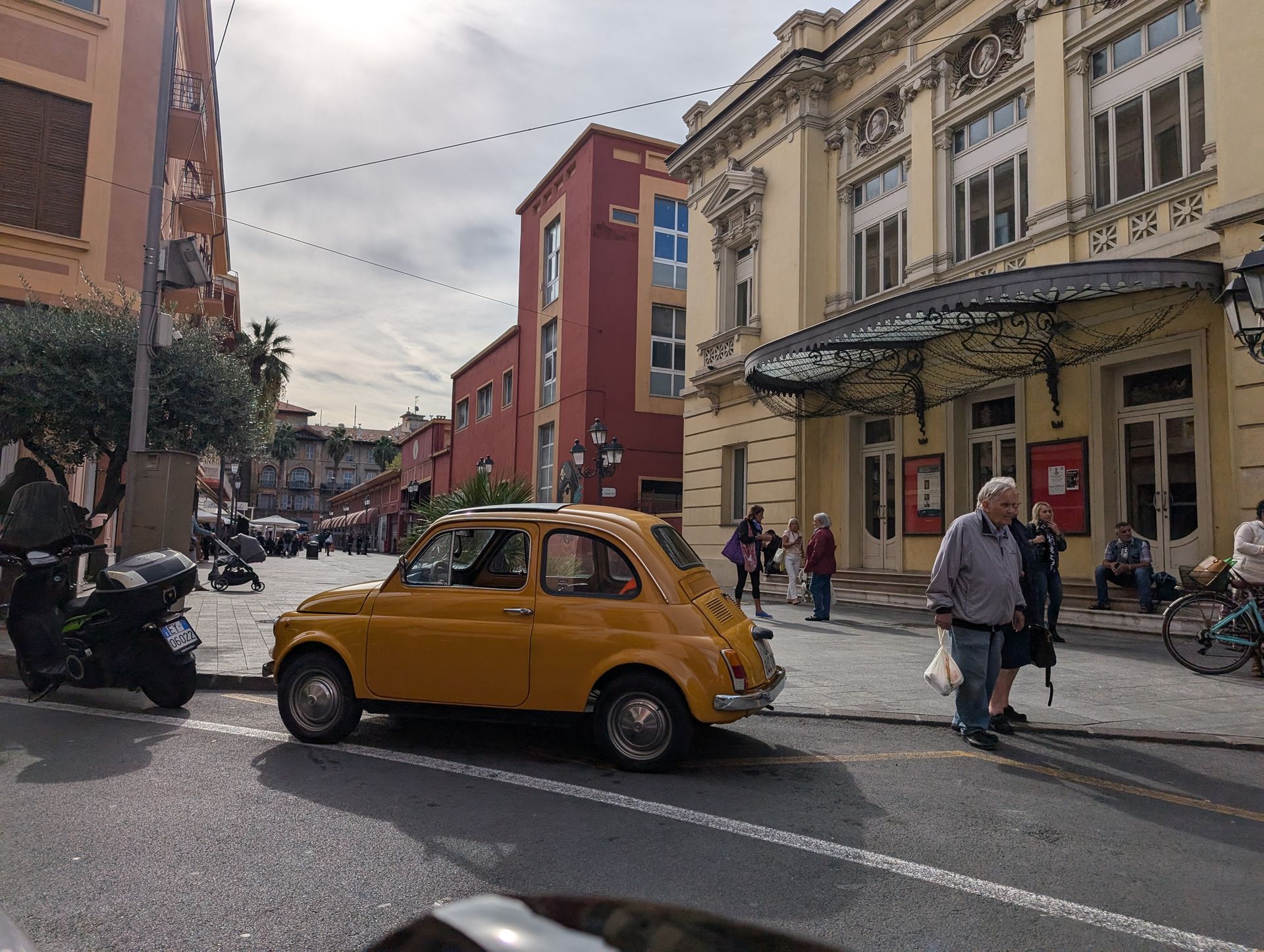 Yellow vintage car on a city street, people walking, a theater with ornate facade in the background.