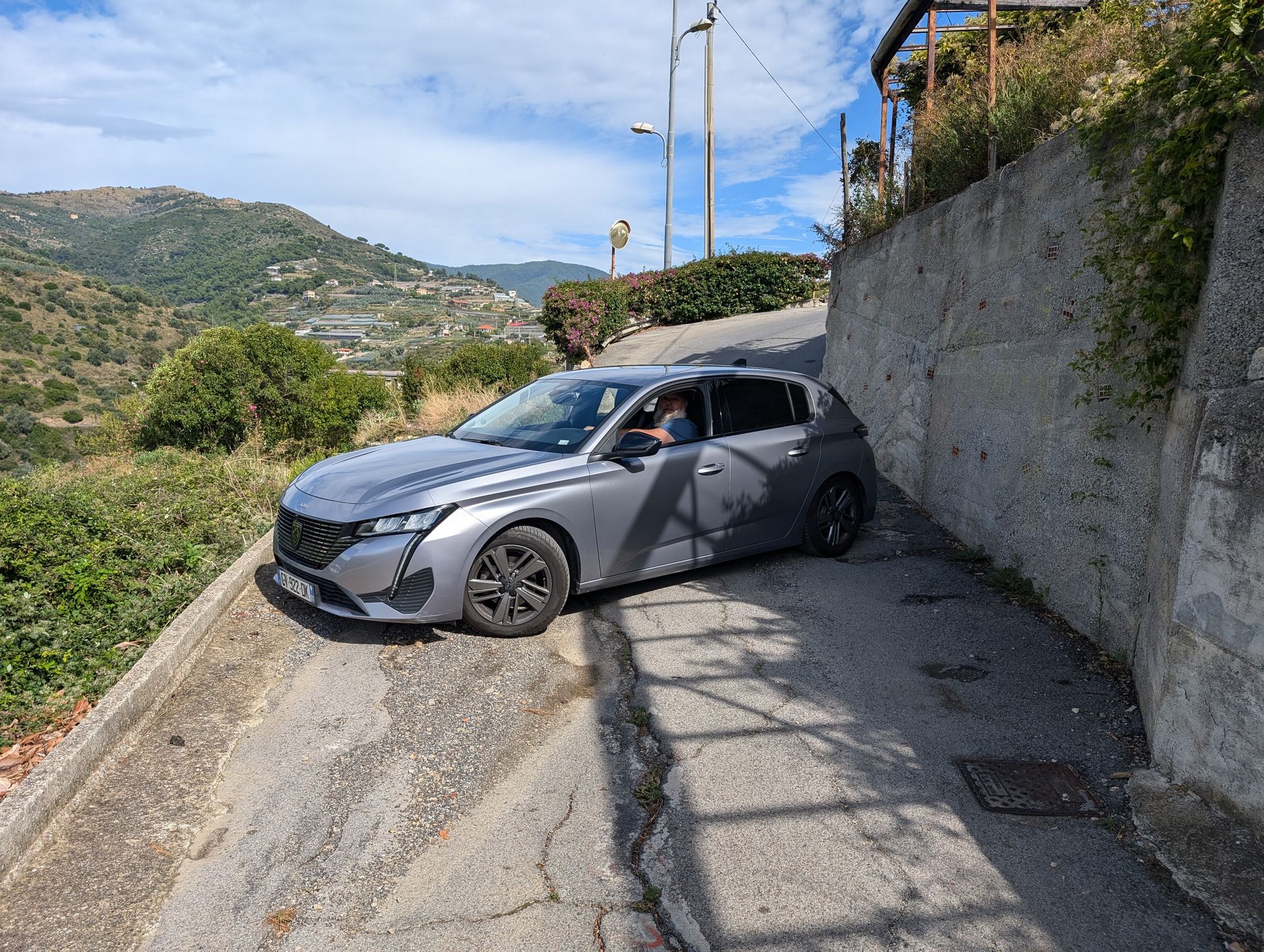 Gray car on a narrow, sloped road with stone walls, mountainous background, and blue sky.