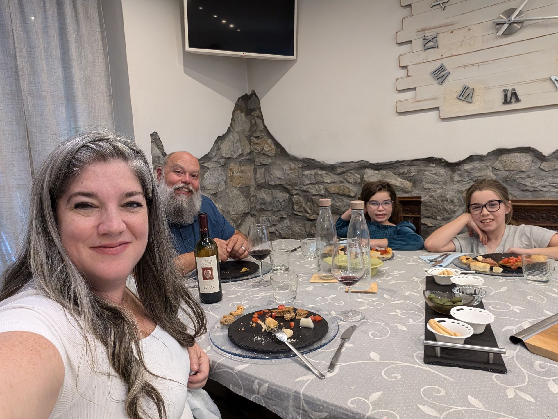Family at a dining table. Woman takes selfie, others seated, food and wine on table. Interior.
