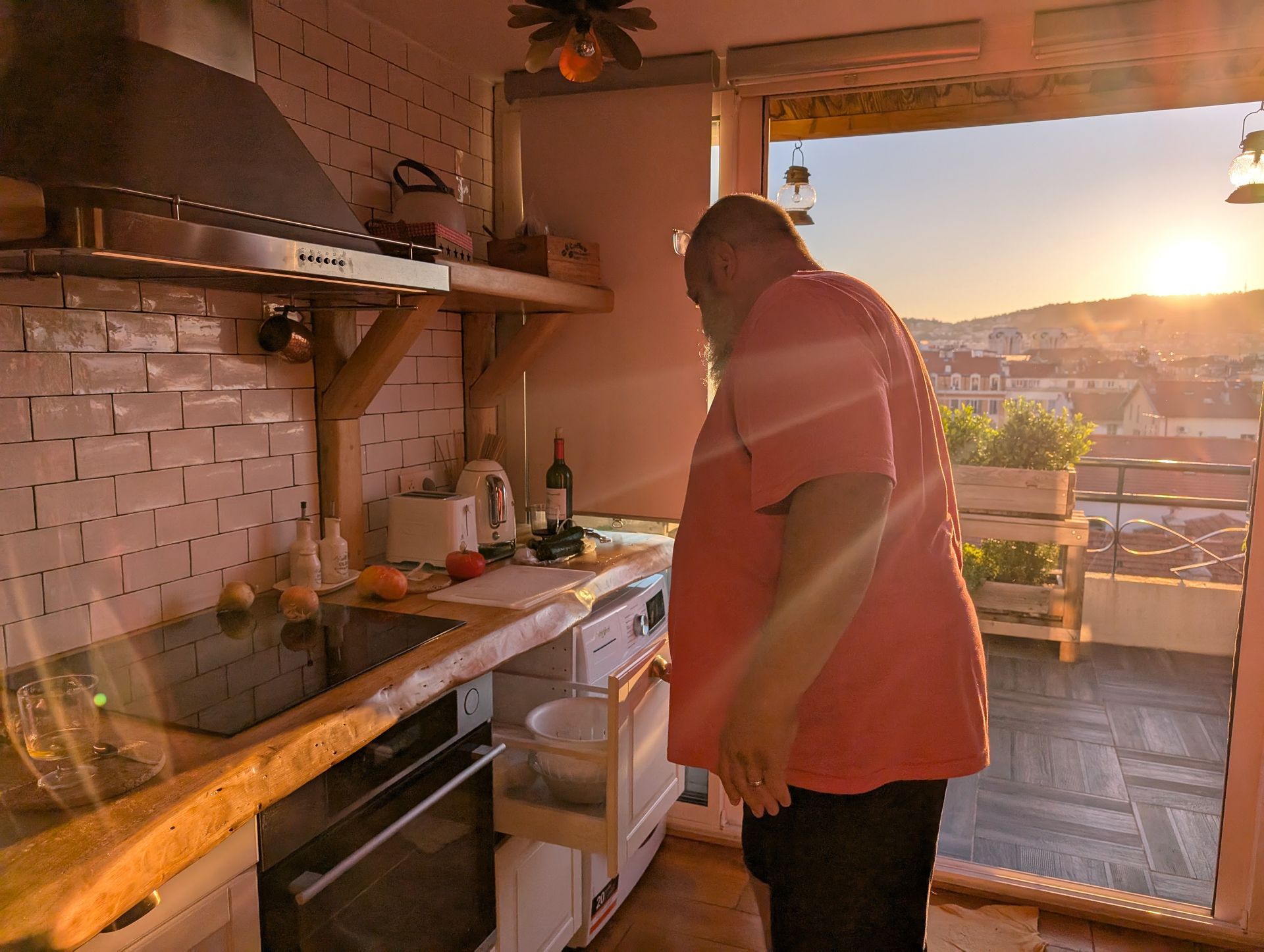 Man cooking in a kitchen, looking out at a sunset over city buildings from an open doorway.