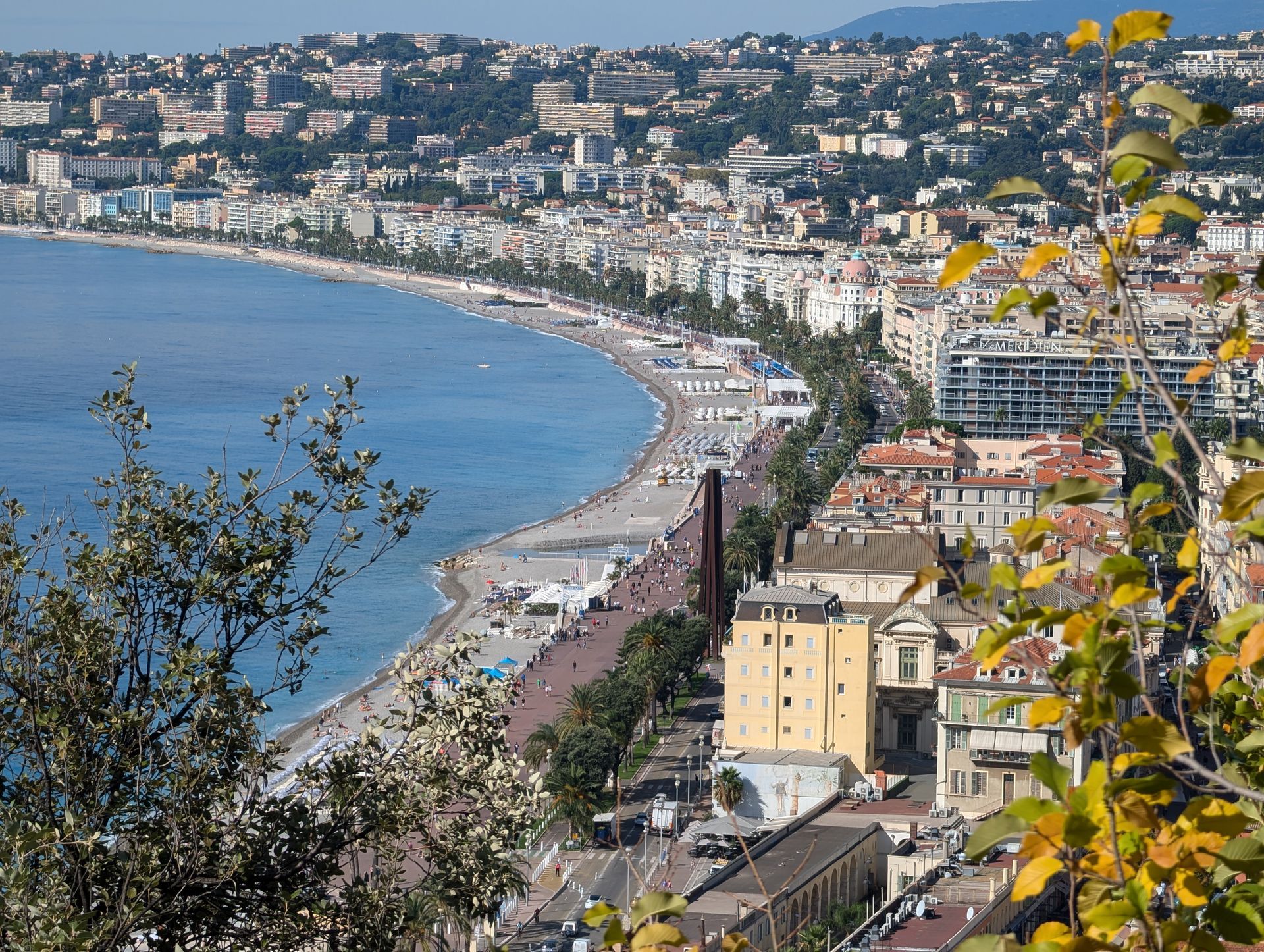 Coastal cityscape with a blue sea, beach, and buildings lining the shore under a clear sky.
