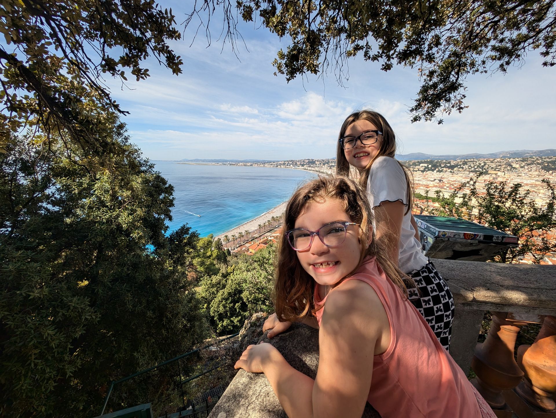 Two girls smile at the camera, overlooking a city and turquoise sea.