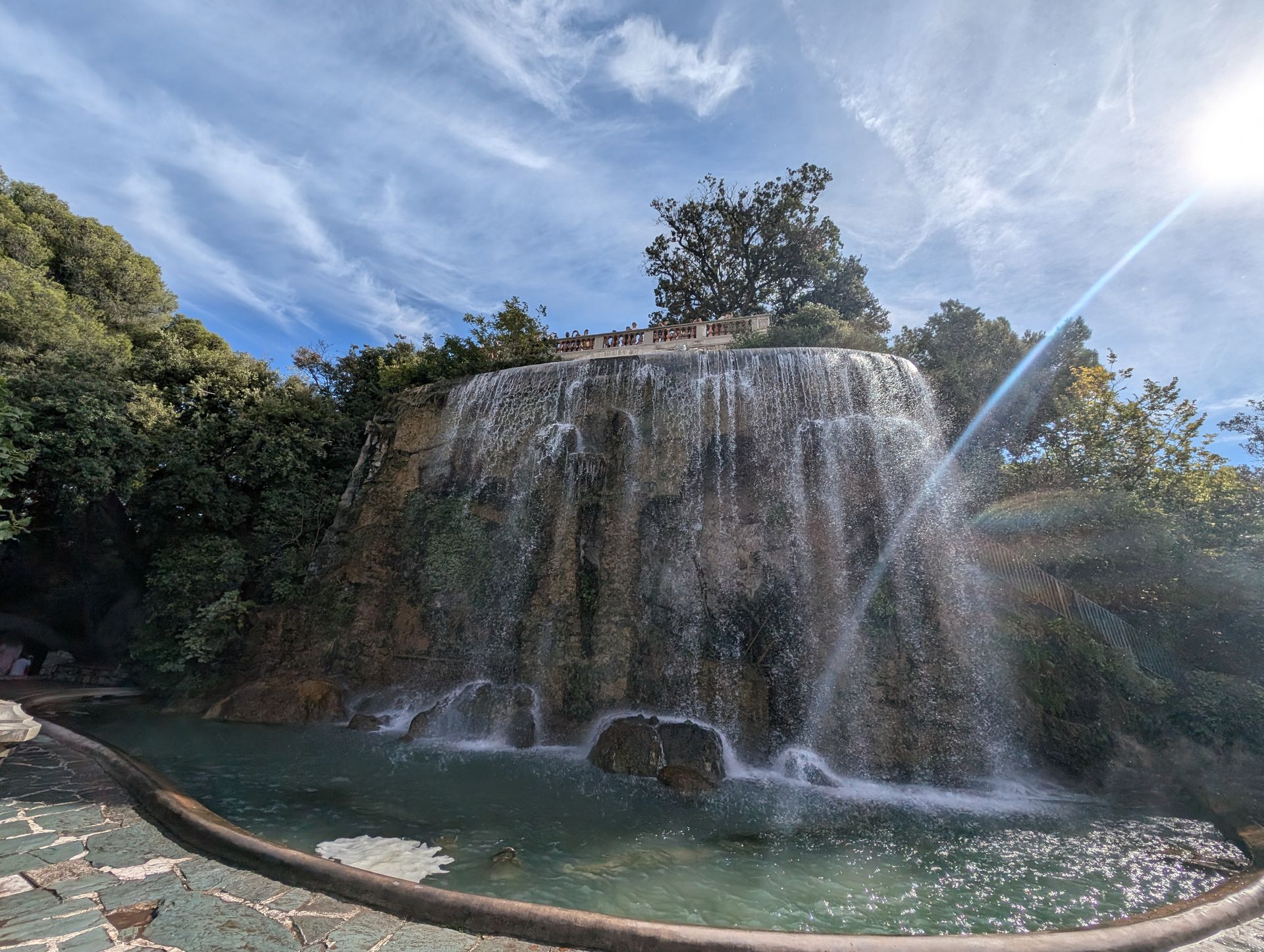 Waterfall cascading into a pond, surrounded by lush greenery, under a blue sky with clouds.