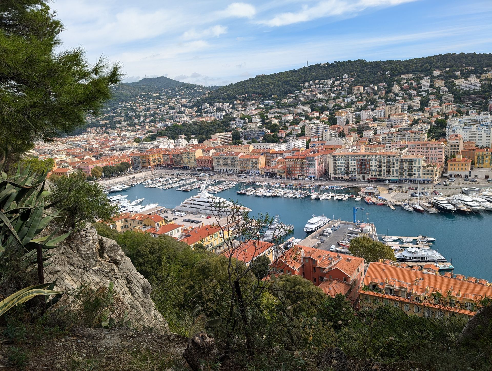 Coastal city harbor view with boats, red rooftops, and green hills.