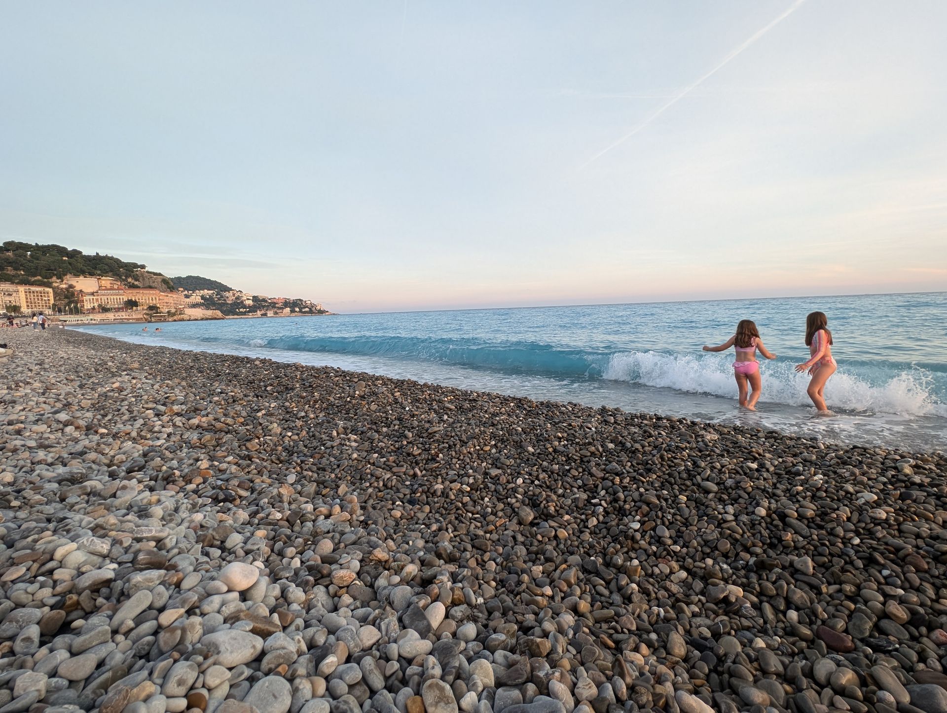 Two children in pink swimsuits playing in the waves on a pebble beach with a town in the distance.