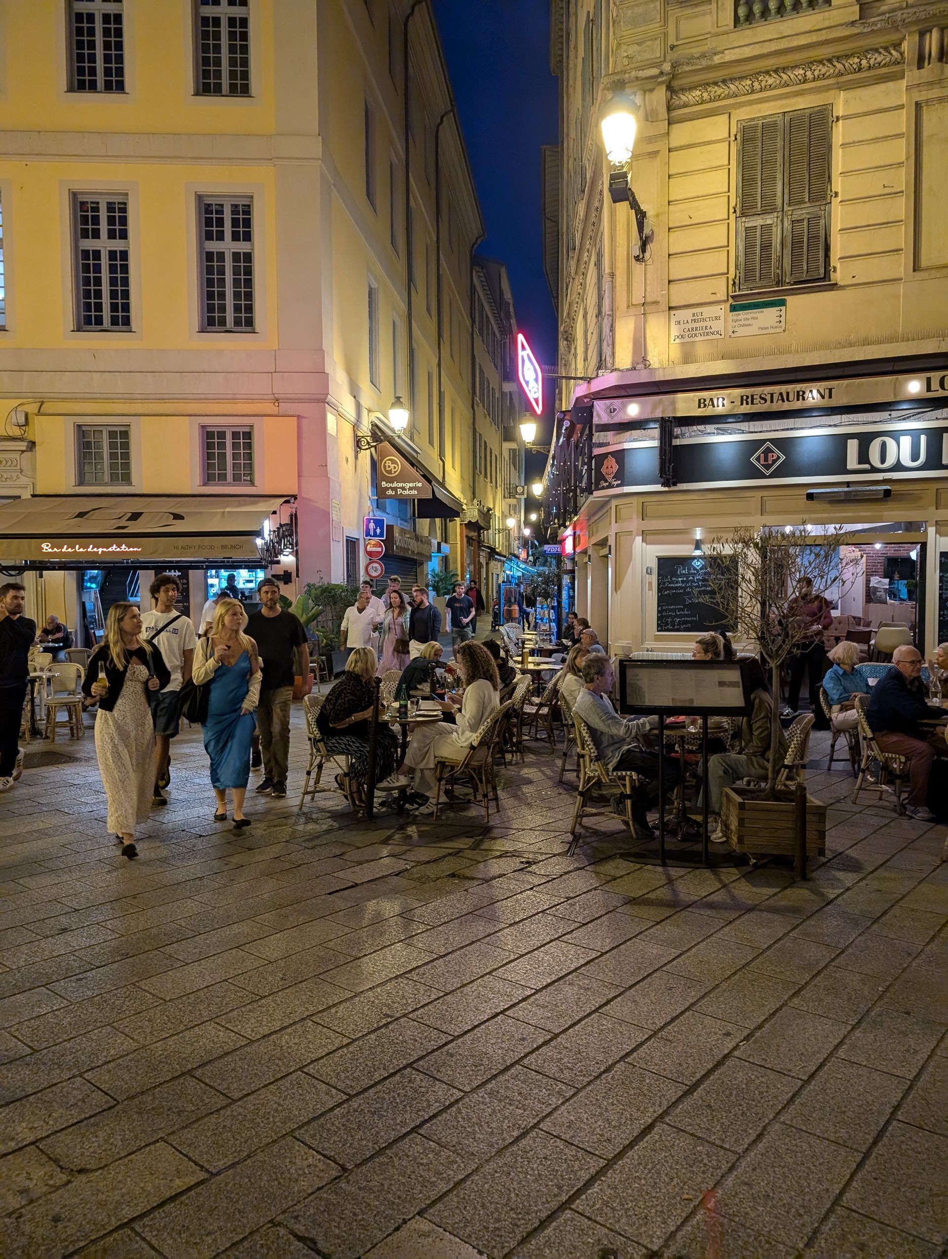 Narrow European street at dusk with people dining and walking past lit restaurants.