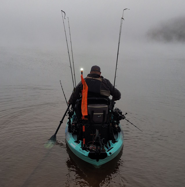 Person fishing from a teal kayak in foggy water, with multiple fishing rods and a light.