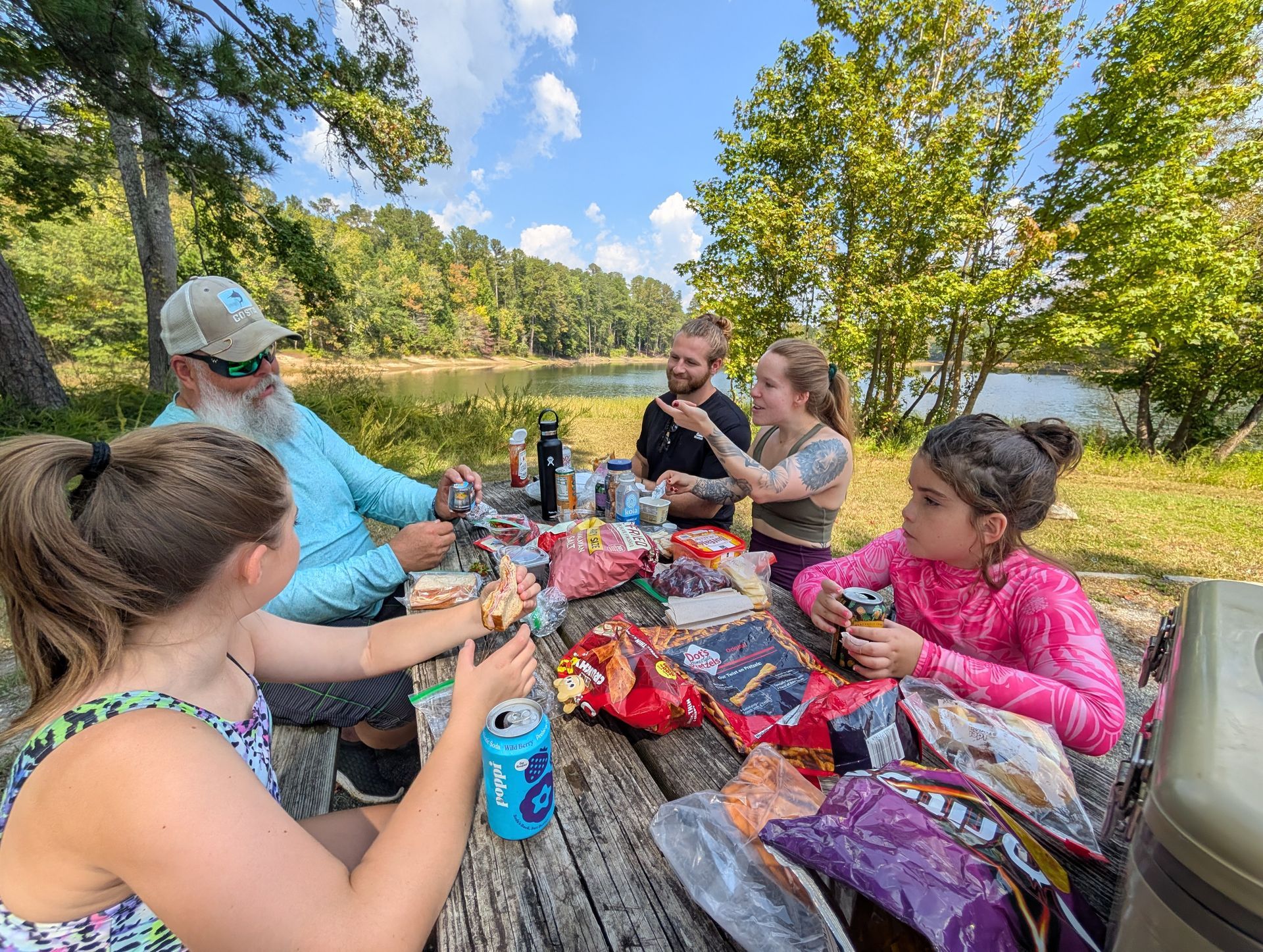 Family picnicking at a lakeside park. People are eating snacks at a wooden table. Sunny day.