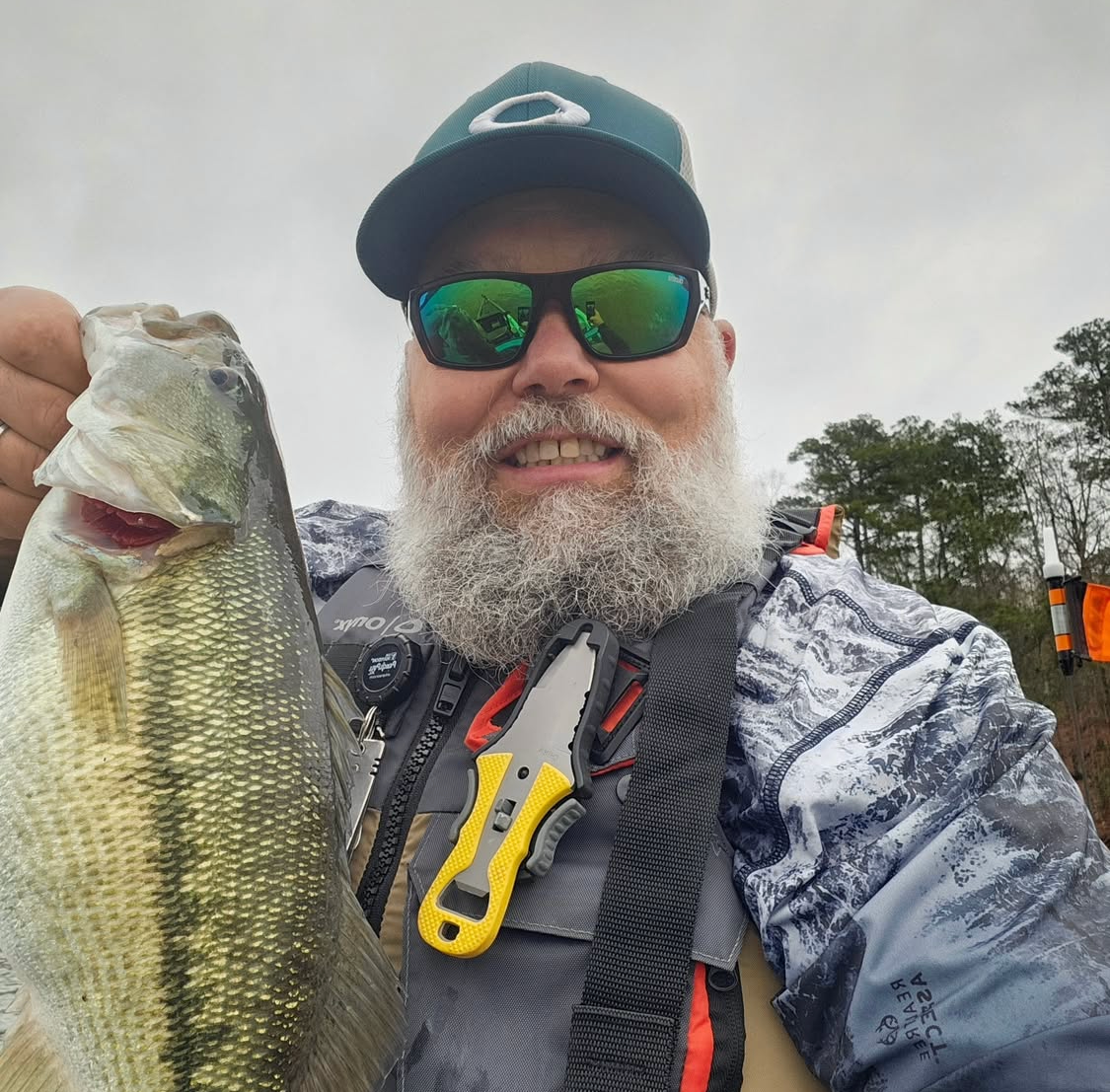 Man holding a large bass fish; outdoors on overcast day. He’s wearing sunglasses, hat, life vest, and smiling.