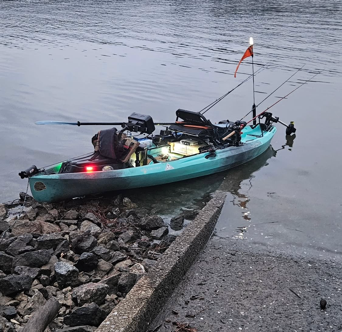 Fishing kayak loaded with gear sits at the edge of a shoreline. A flag and multiple fishing rods are visible.