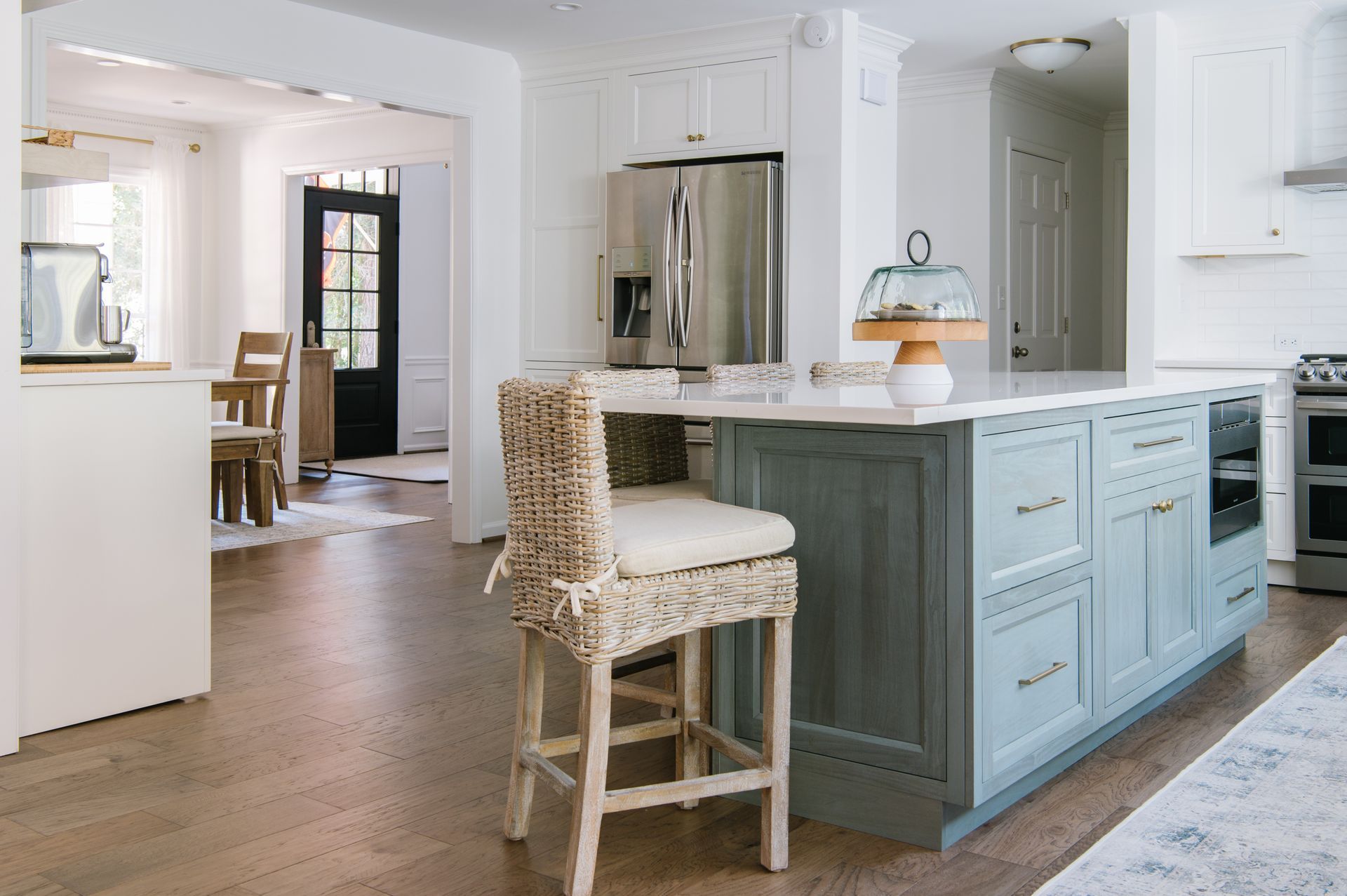 Kitchen with blue island, stainless steel fridge, and rattan bar stool. Wooden floor and white walls.