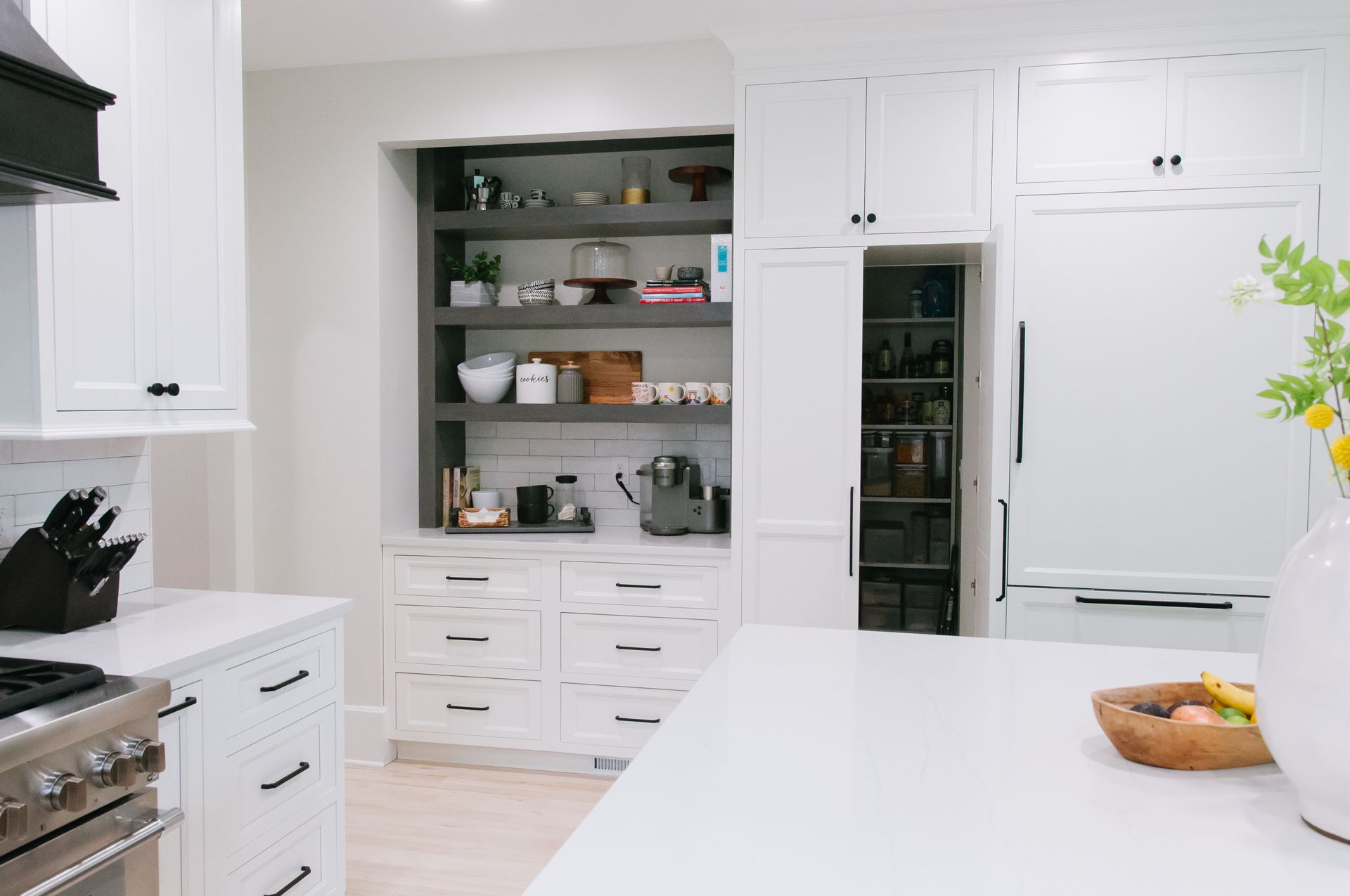 White kitchen with built-in shelves, drawers, and pantry. A large island and flowers are in view.