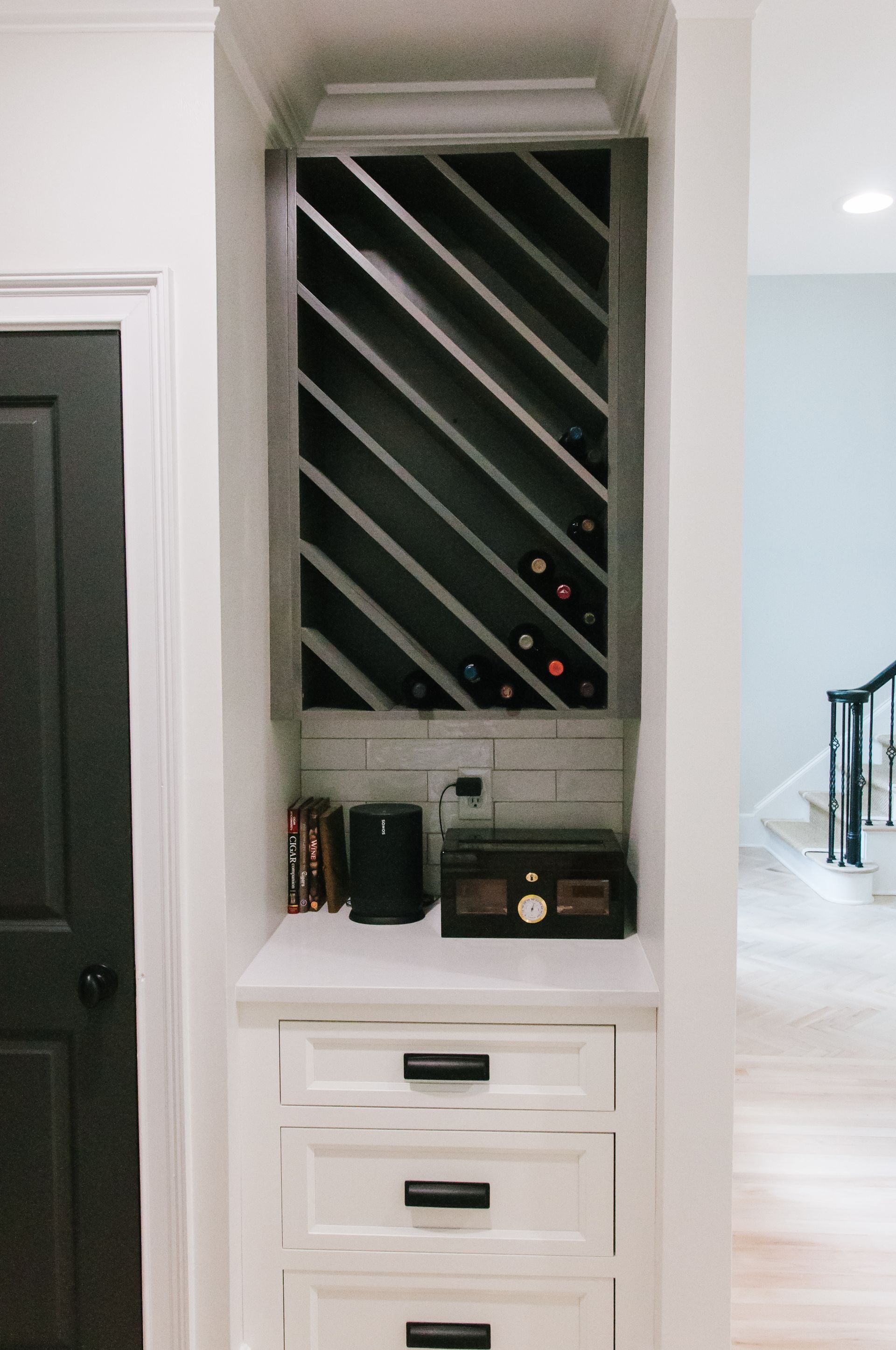 Built-in wine rack with bottles, drawers, and counter in a white-walled alcove. Black door on the left.