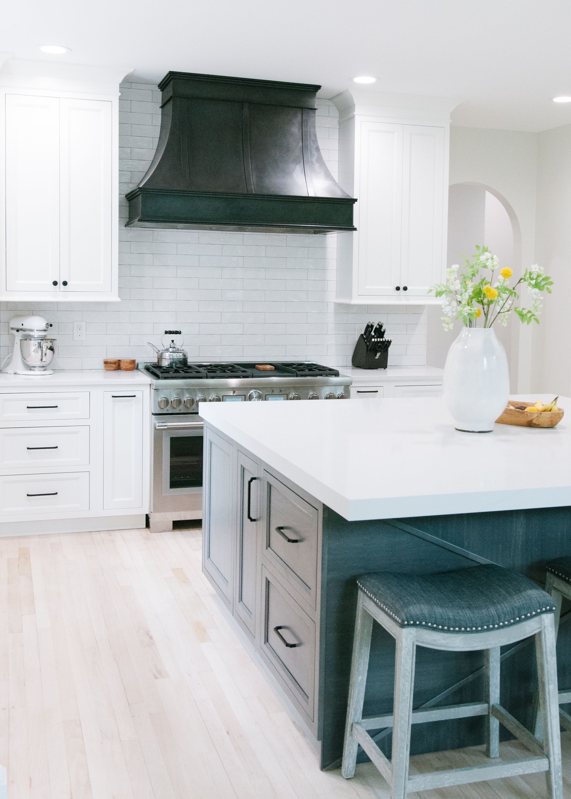 A modern kitchen with white cabinets, blue island, stainless steel stove, and a dark range hood.