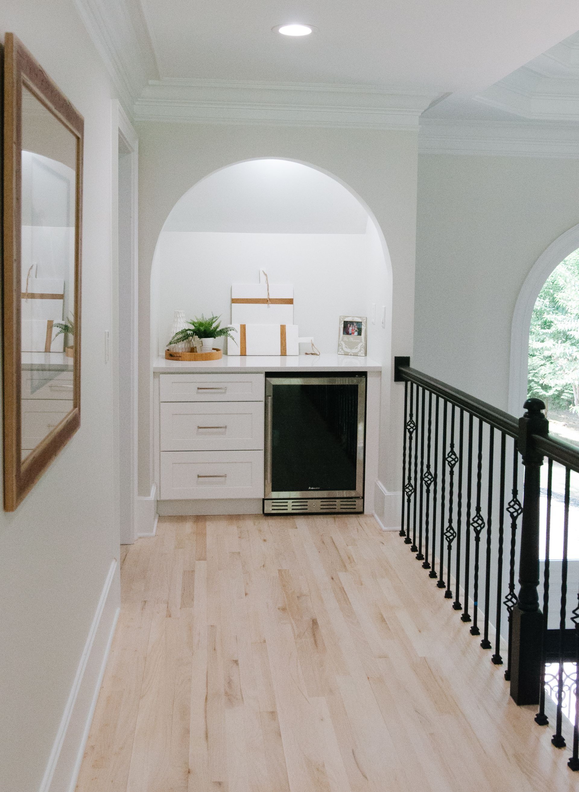 Hallway with arched alcove bar area; white cabinetry, mini-fridge, light wood floors, black railing.