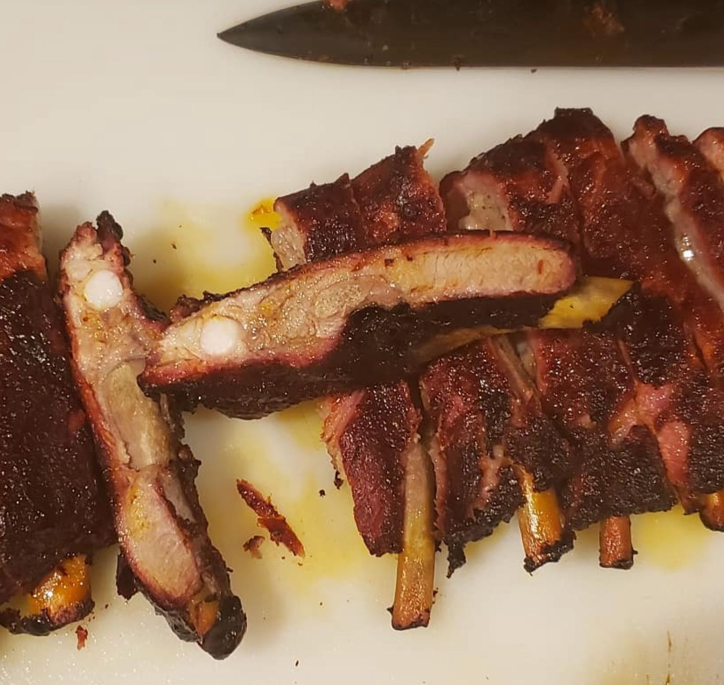 Sliced, smoked ribs on a white cutting board, knife in the background. Brown, charred exterior with pink interior.