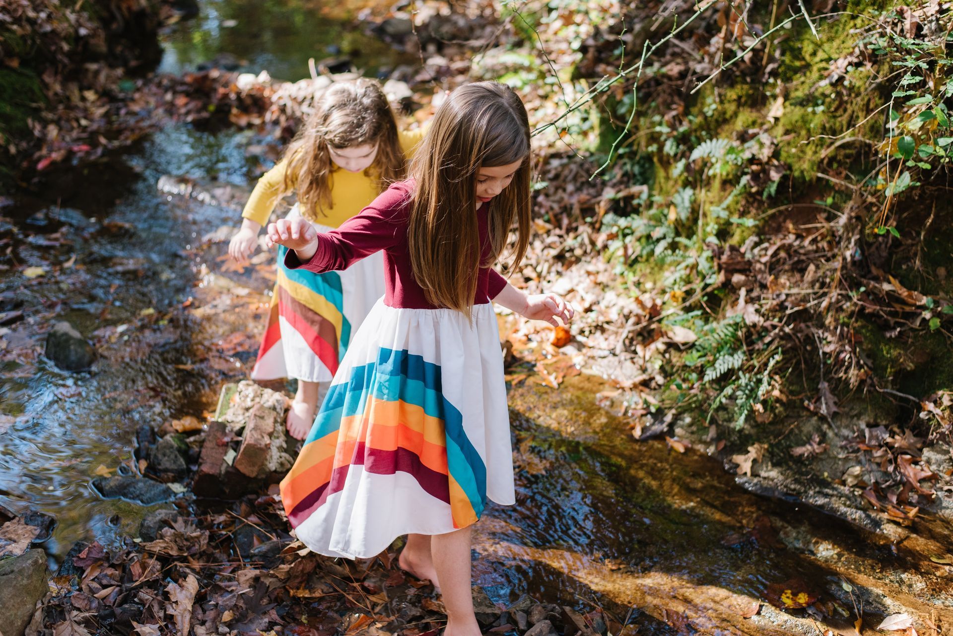 Two children wearing rainbow skirts walk across a shallow stream.