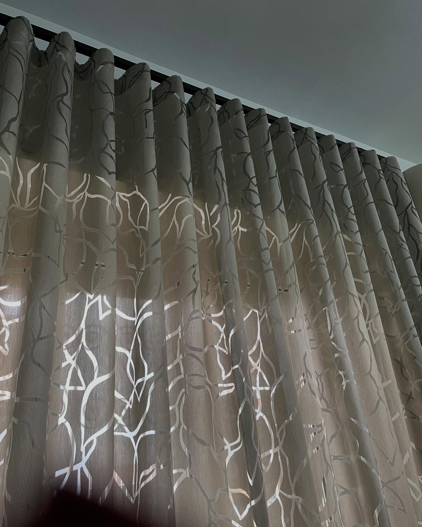 Tende trasparenti beige con motivo a foglie bianche, appese a un'asta nera.