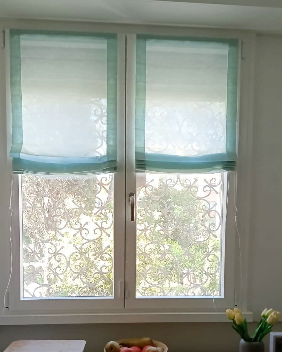 Finestra a due vetri con tende romane blu-verdi e pellicola decorativa interna; sul davanzale c'è un vaso di fiori.