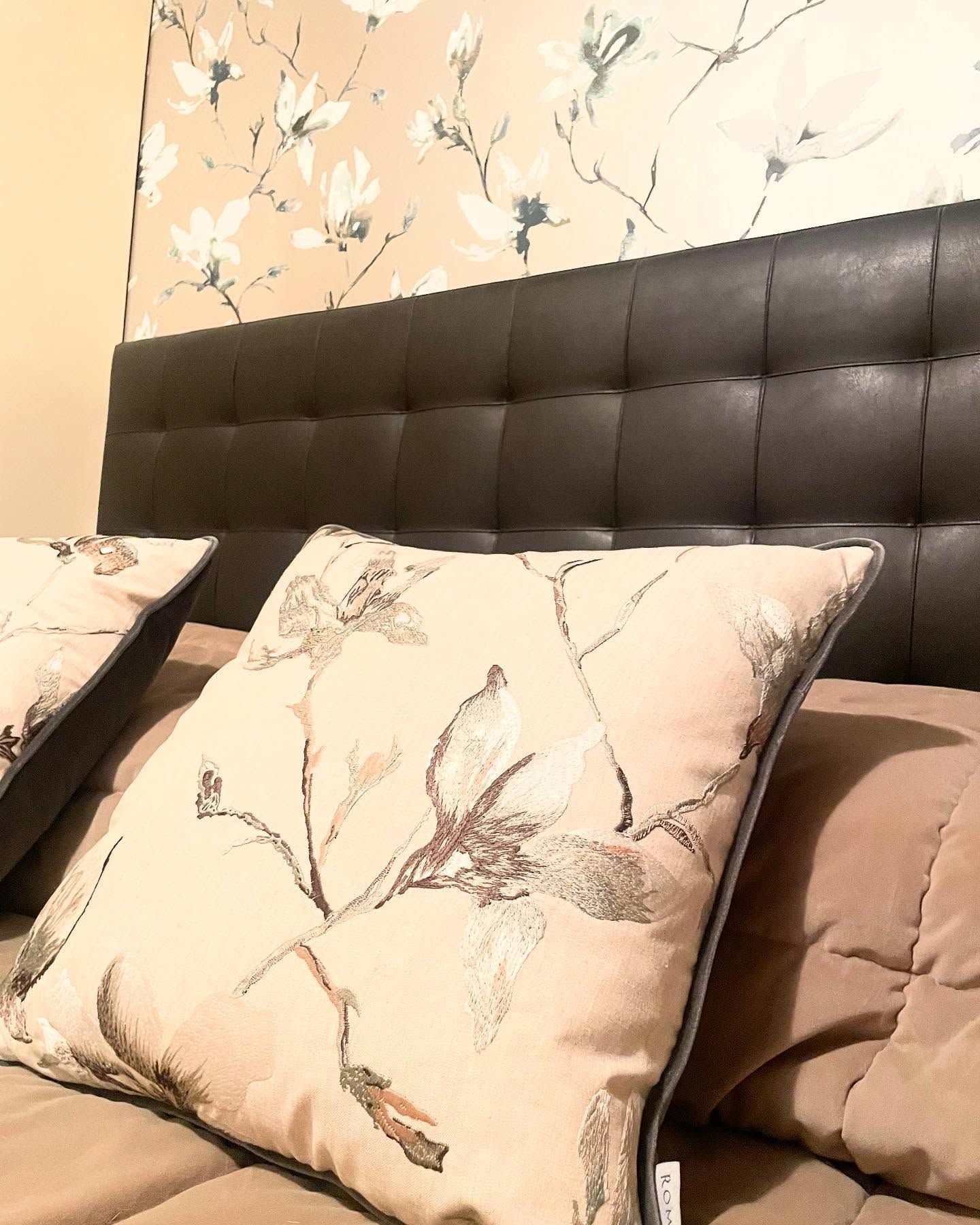 Letto con testiera nera, cuscini floreali e carta da parati con fiori bianchi.