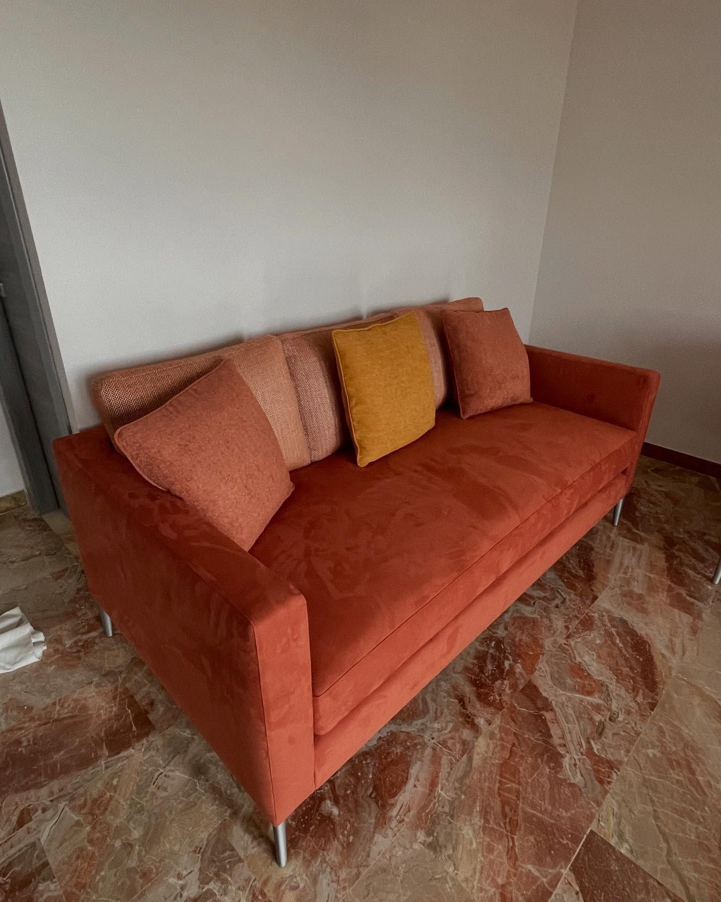 Divano arancione con cuscini contro una parete bianca. Il divano è appoggiato su un pavimento decorato.
