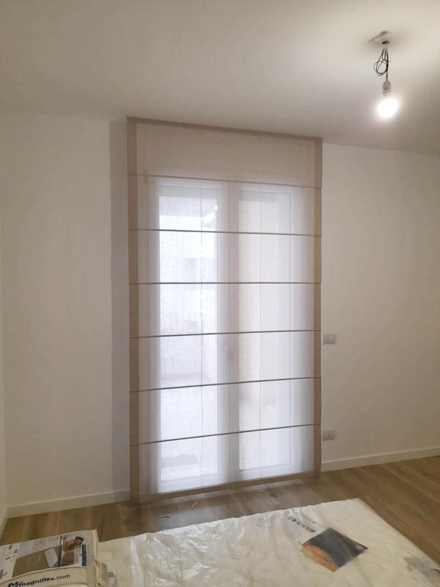 Una tenda romana bianca copre una finestra. La tenda è incorniciata da profili beige.