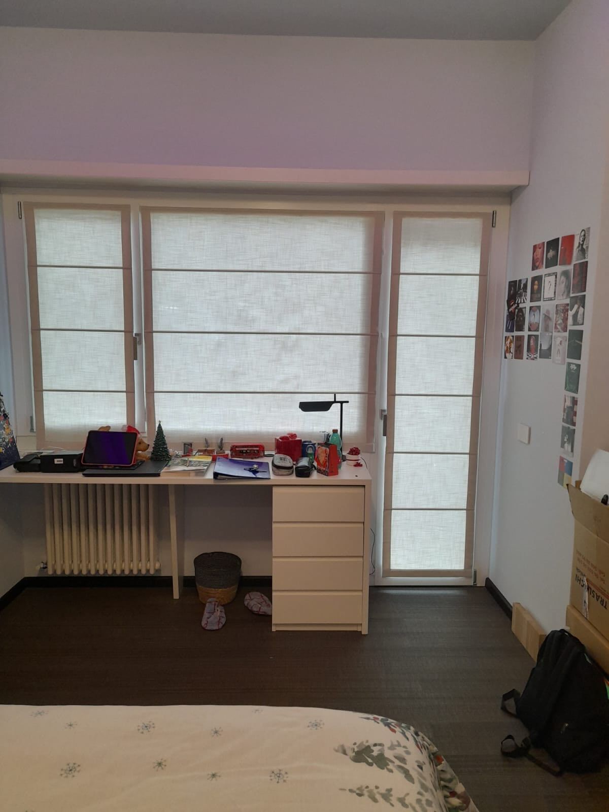 Una camera da letto con scrivania, finestra e porta. Scrivania bianca con cassetti, termosifone e tende alla romana.