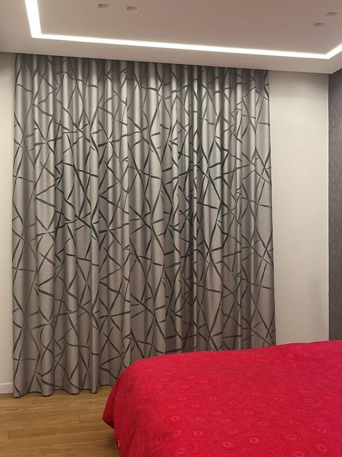 Tende grigie con motivi geometrici neri in una camera da letto con copriletto rosso e pavimento in legno.