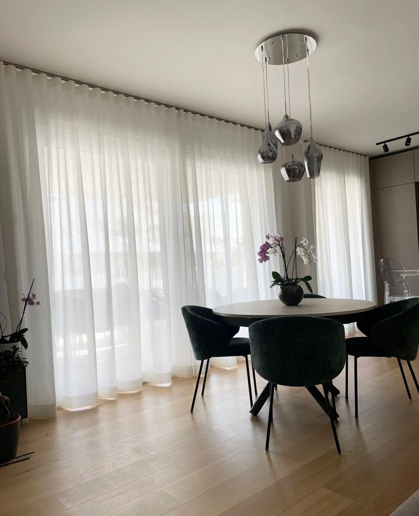 Zona pranzo con tavolo rotondo, sedie verdi, tende bianche trasparenti e lampadario cromato.